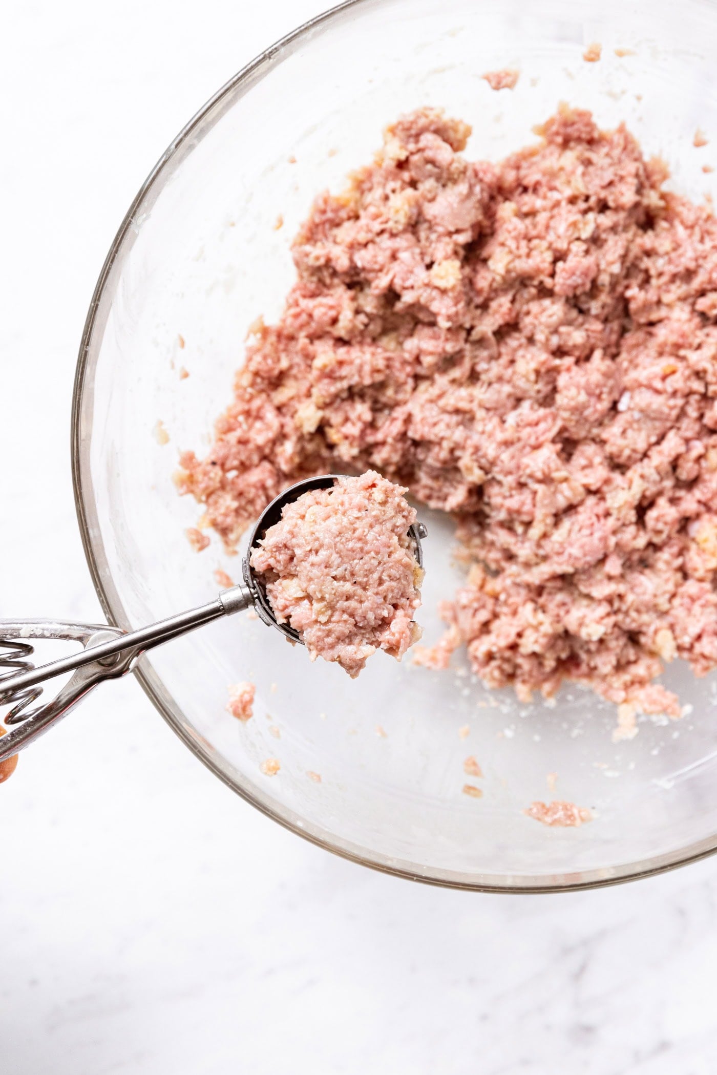 Glass bowl sitting on marble surface with ice cream scoop scooping out a small serving of ground meat for meatballs.