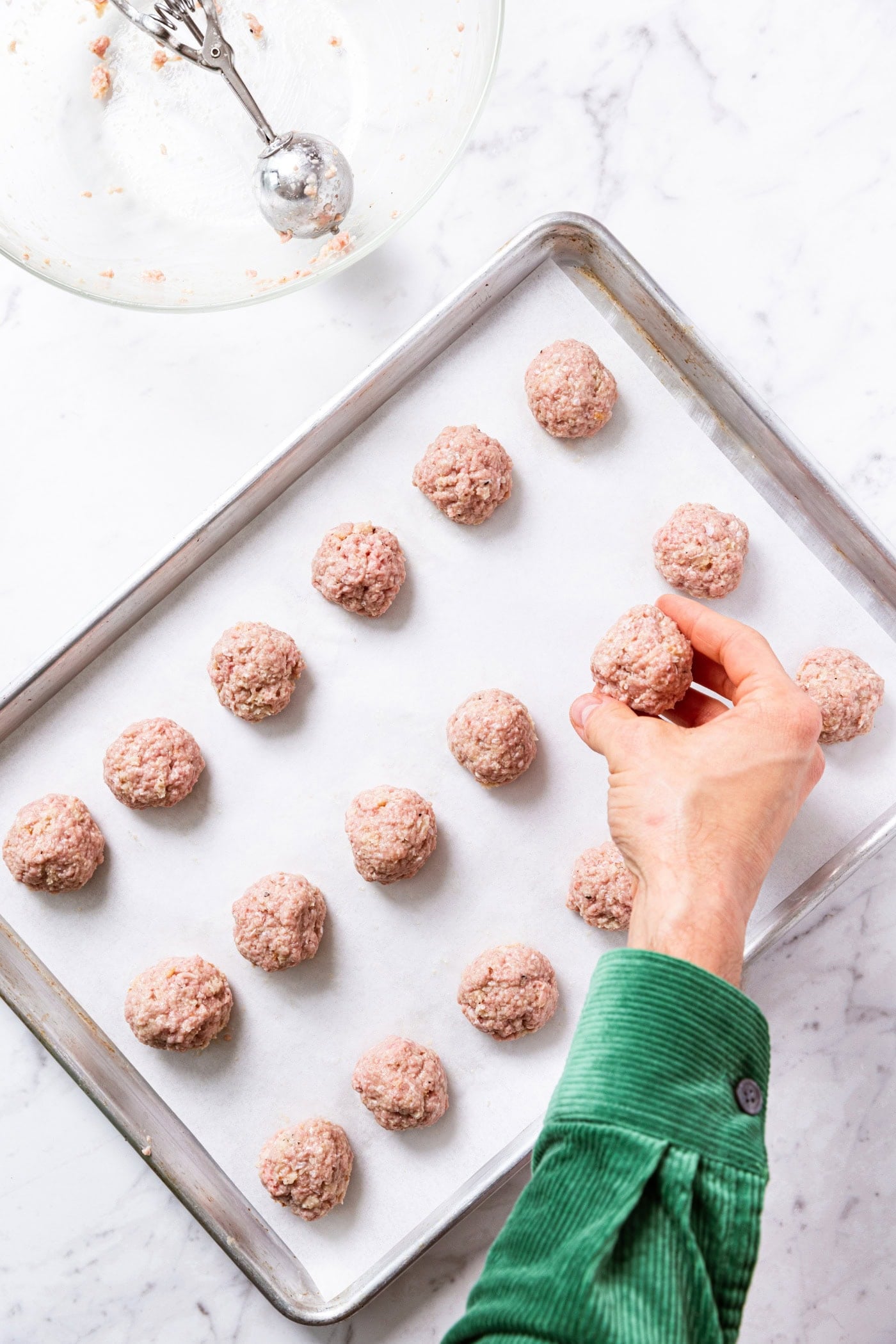 Hand placing rolled meatball on parchment-lined metal baking sheet on marble surface with empty glass bowl and ice cream scoop in bowl in upper corner.