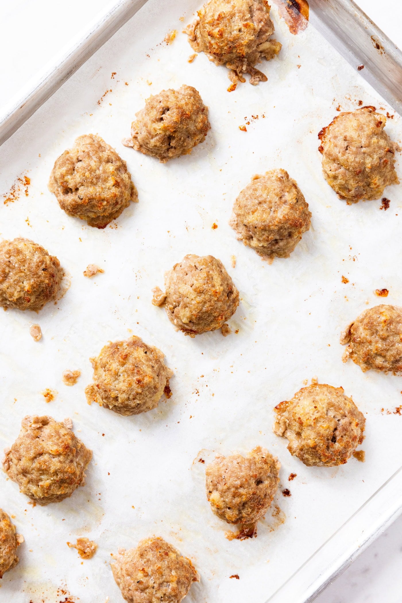 Parchment-lined baking sheet lined with baked meatballs with bits of baked pieces all around sitting on white marble surface.