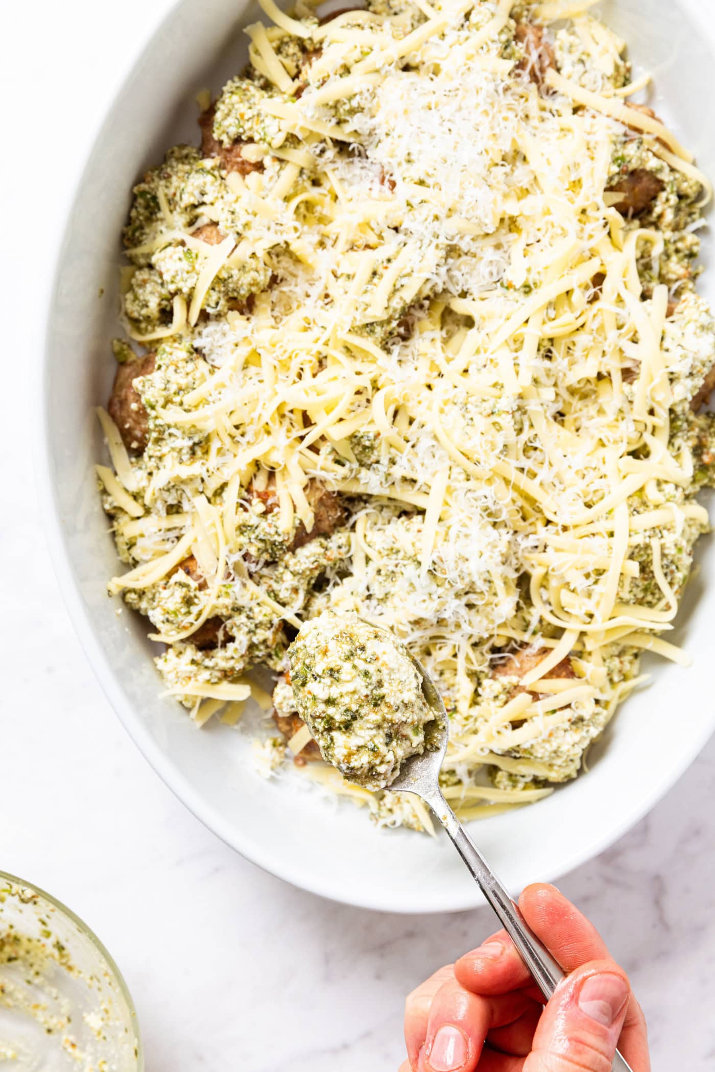 Spoon placing pesto mixture on top of baked meatballs in oval baking dish with Parmesan cheese sprinkled all over the top.