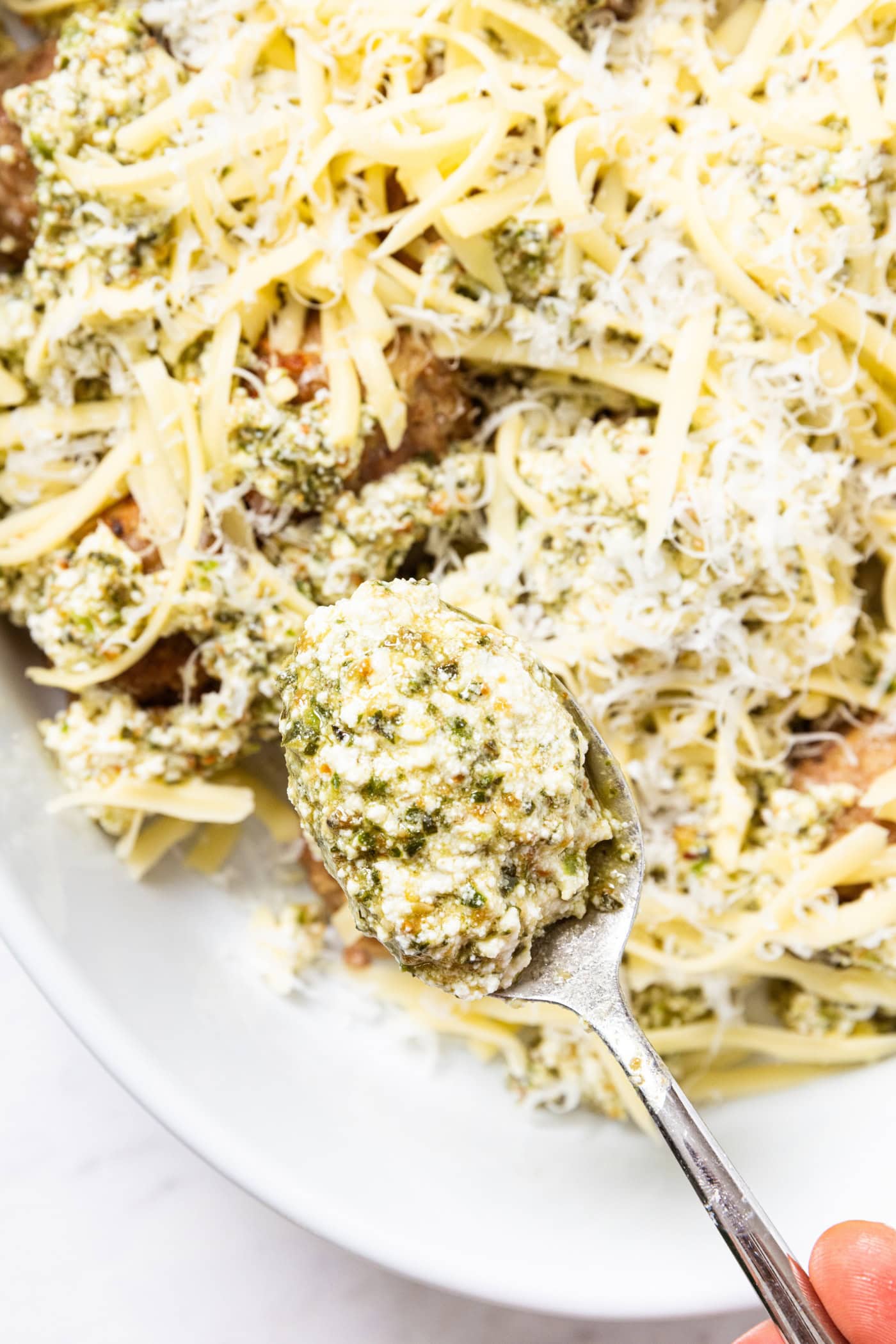 Spoon holding pesto mixture spooning on top of meatballs after coming out of oven along with Parmesan cheese all in white oval baking dish.