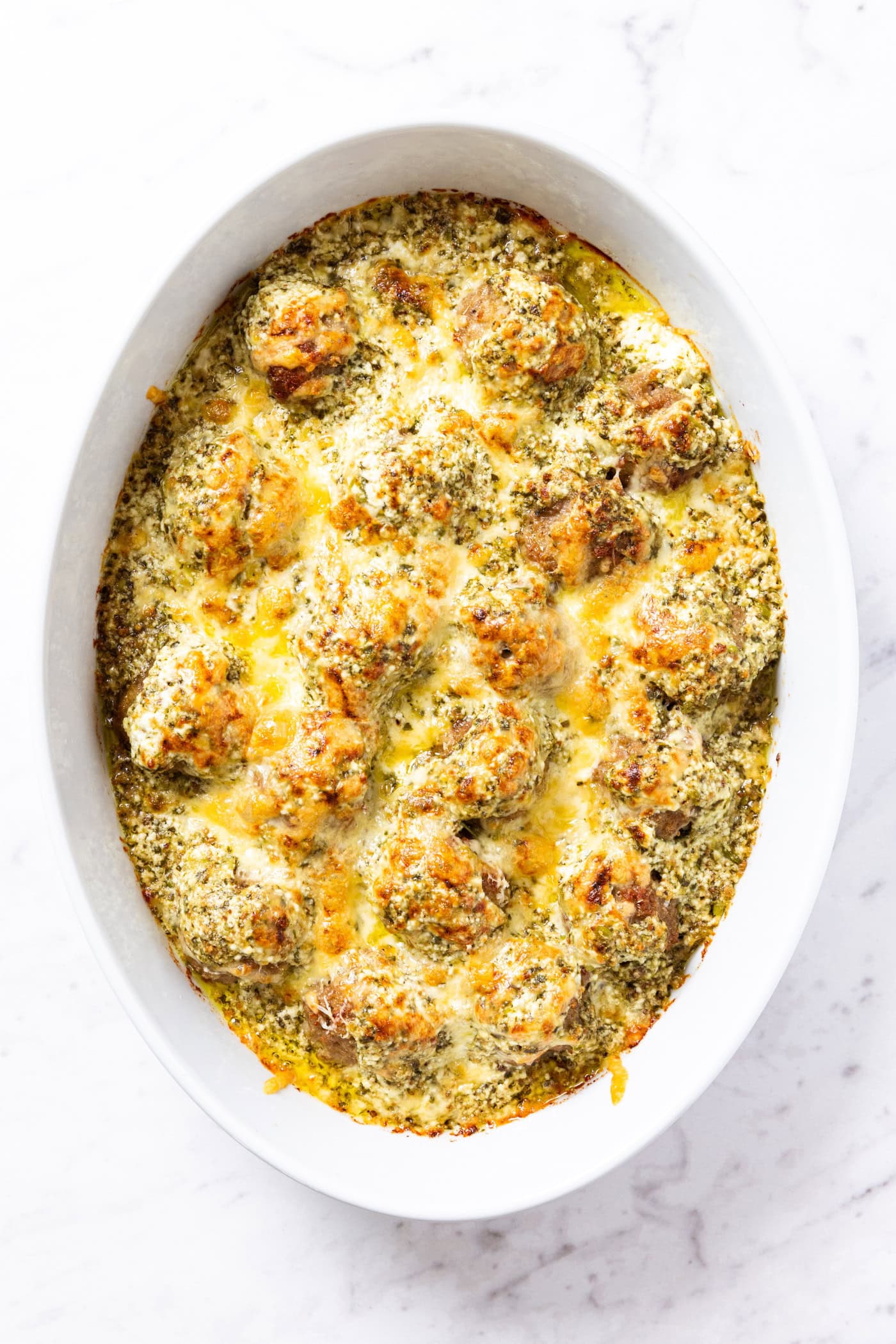 Oval baking dish on marble surface with baked meatballs topped with Parmesan cheese all over the top showing browned top.