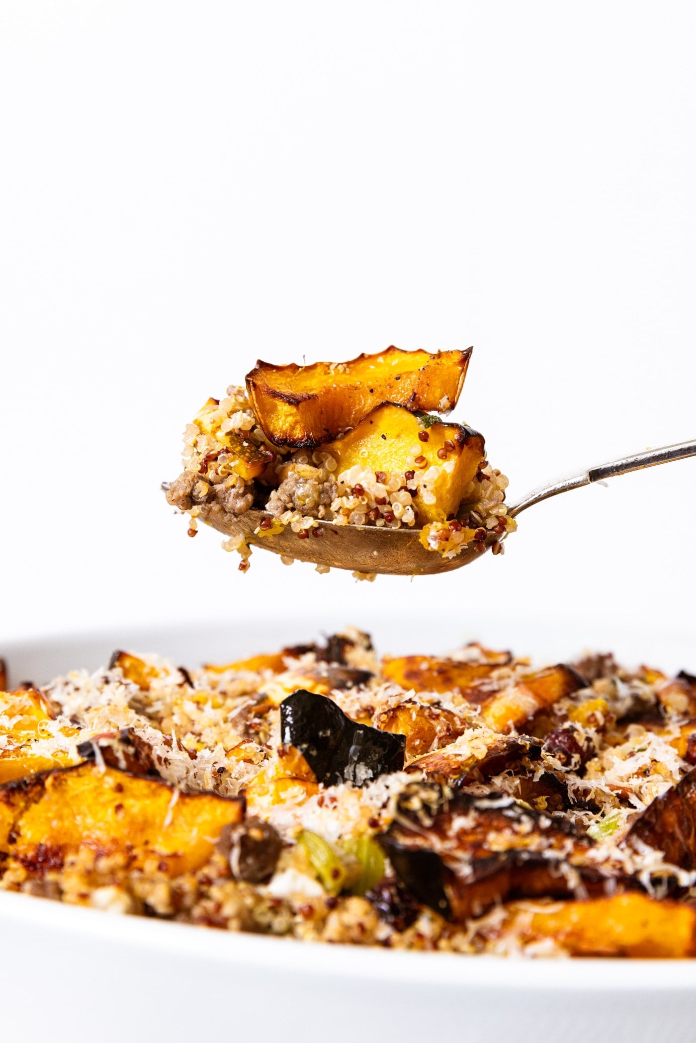 Spoon holding serving of stuffed acorn squash stuffing with rest of casserole still in white baking dish below all with white background.