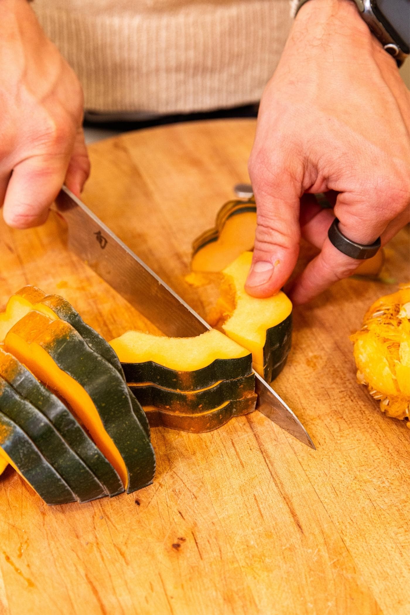 Hand holding knife cutting pieces of acorn squash in quarters on wood cutting board.