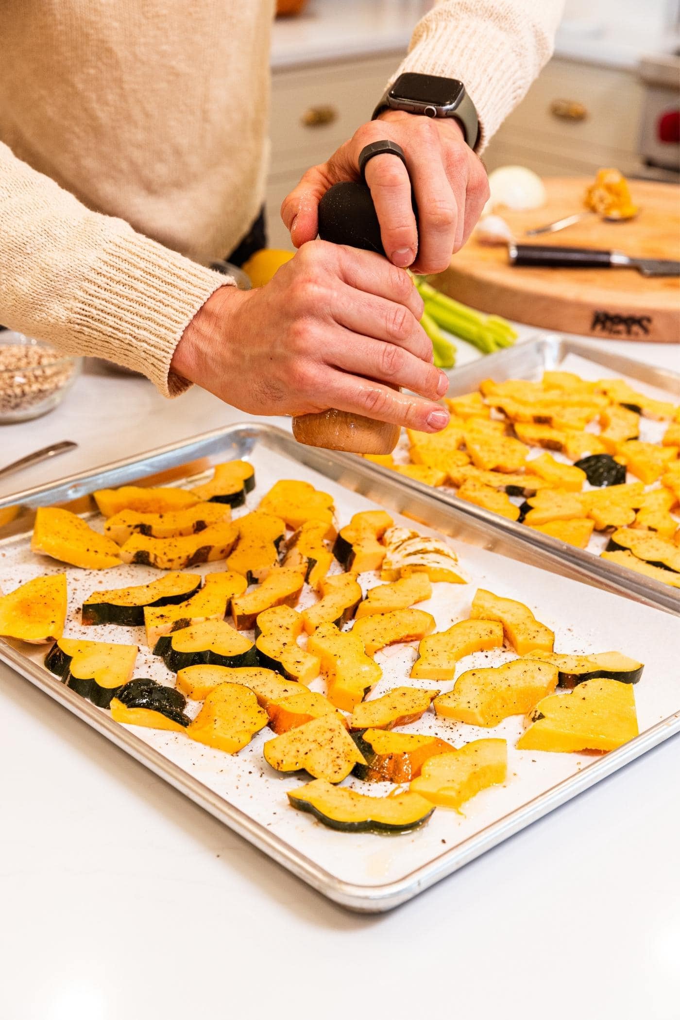 Hand using a pepper grinder to place black pepper all over the top of oiled and salted pieces of acorn squash on a parchment-lined baking sheet on white countertop.