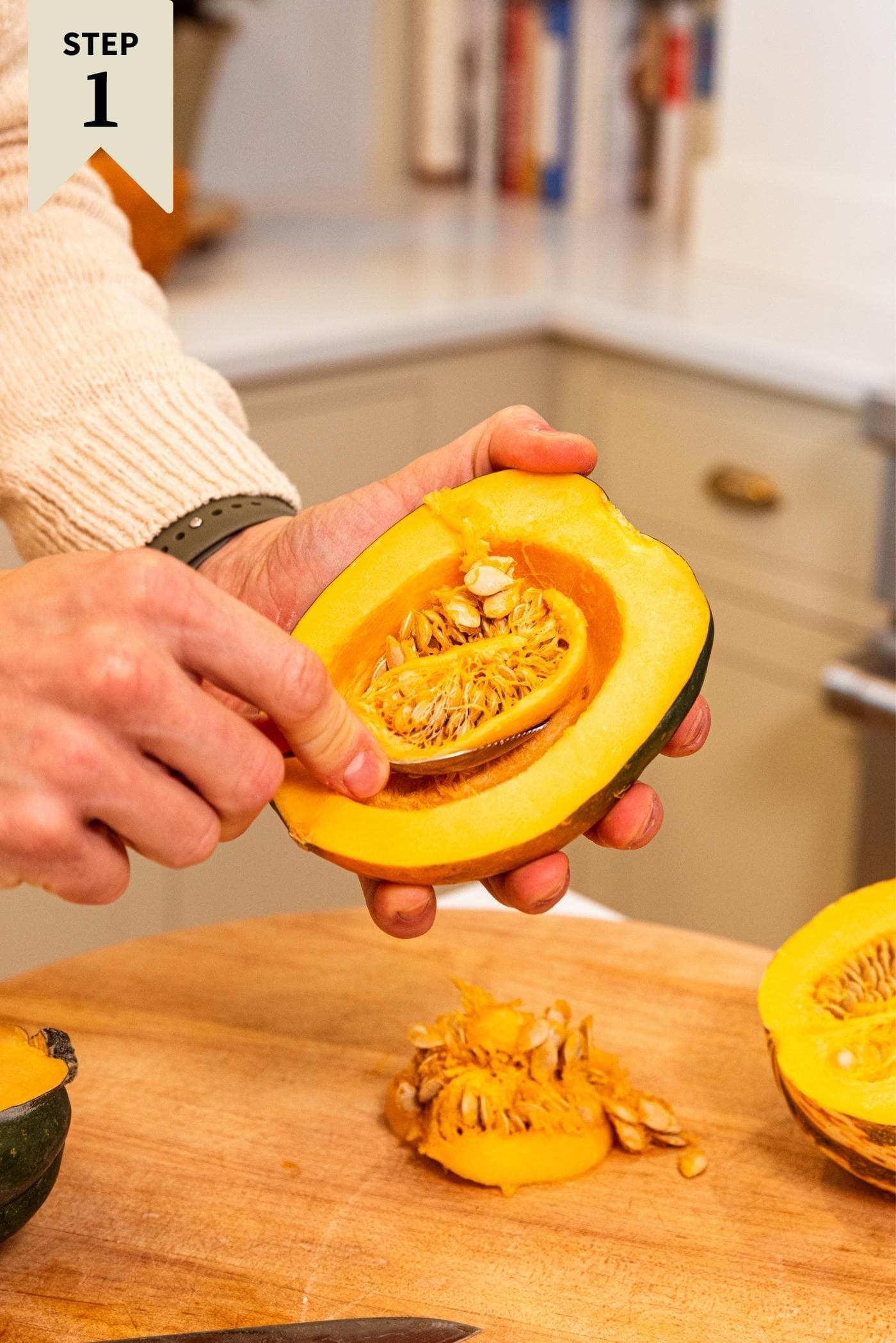 Hand scooping out middle of acorn squash with green skin and placing the seeds on wooden cutting board below.