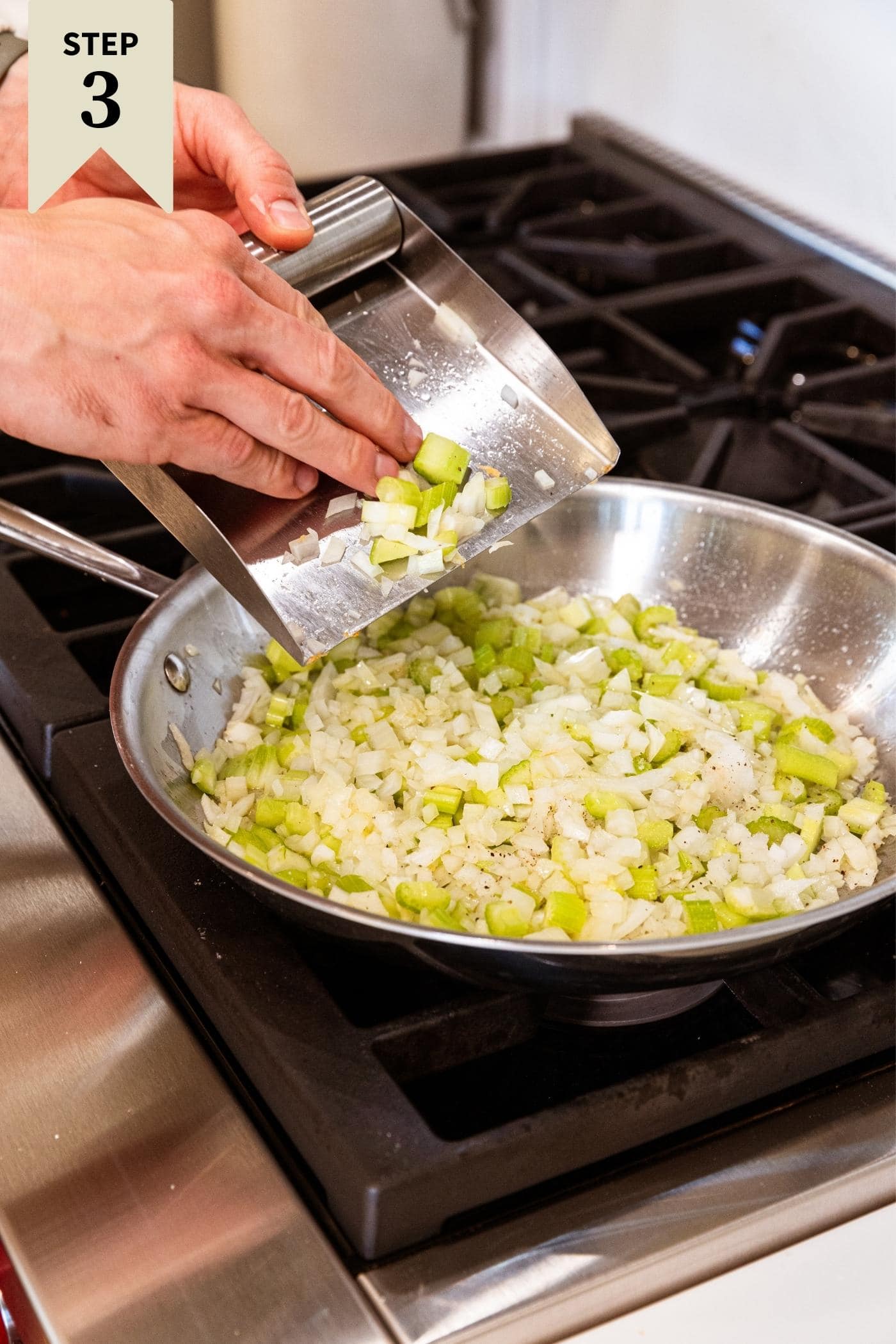 Hand scooping pieces of celery and onion in metal skillet on stovetop with oil in the bottom.