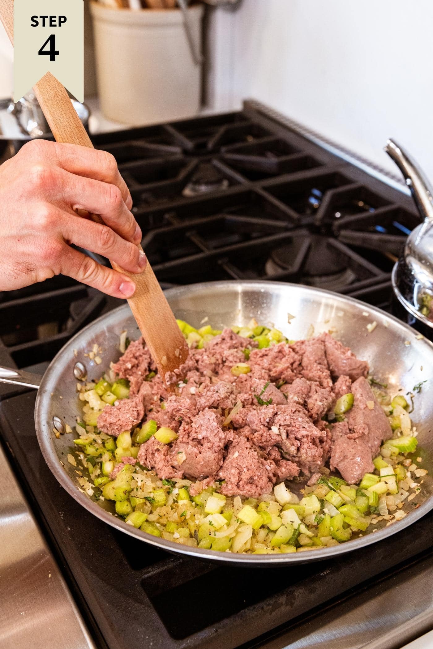 Hand holding wooden spatula mixing together pieces of raw sausage into diced onion and celery mixture cooking in skillet on stovetop.