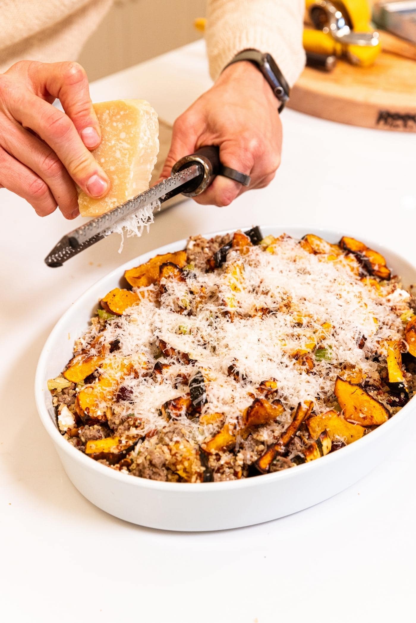 Hand grating Parmesan cheese over the top of a white baking dish filled with squash and sausage stuffing mixture on white countertop.