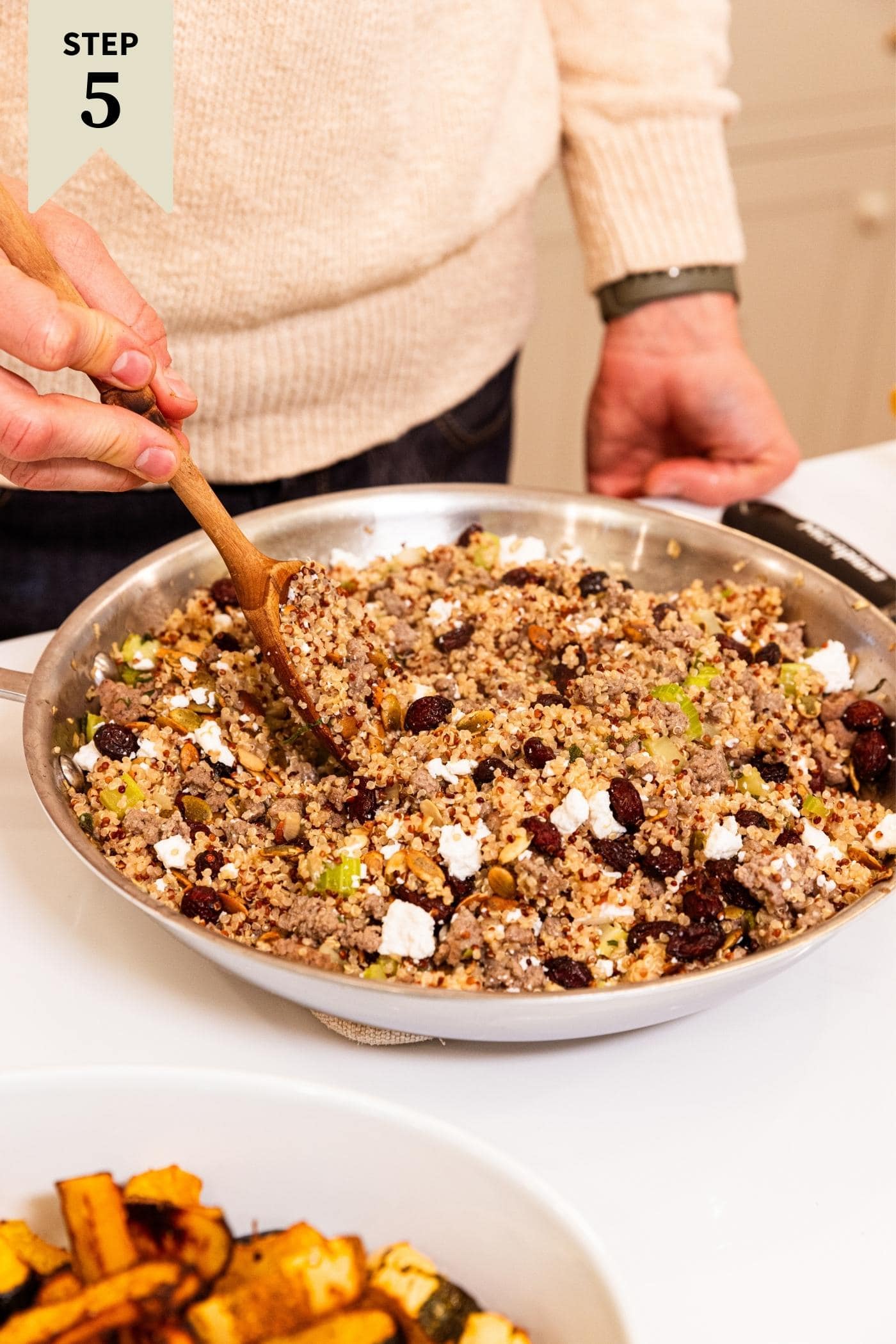 Hand stirring together mixture of quinoa, cranberries, feta, and onion and celery mixture in metal skillet on white countertop with some slices of squash to the side.