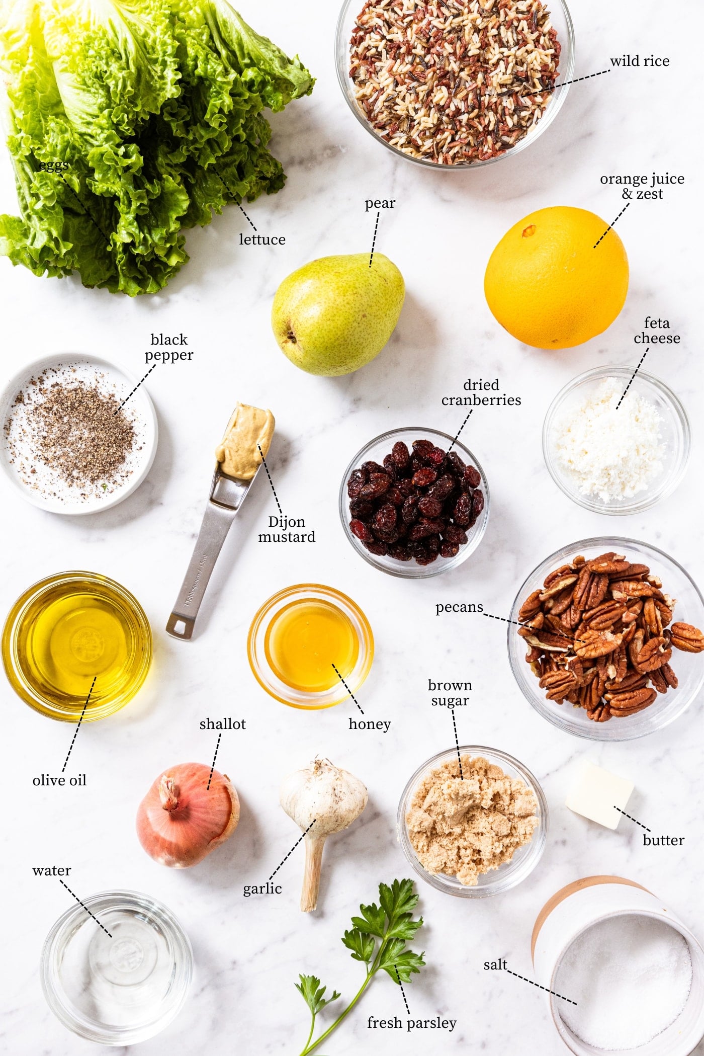 White marble surface filled with all of the ingredients needed to make a wild rice and pear salad including lettuce, spices, nuts, wild rice, olive oil, and more.