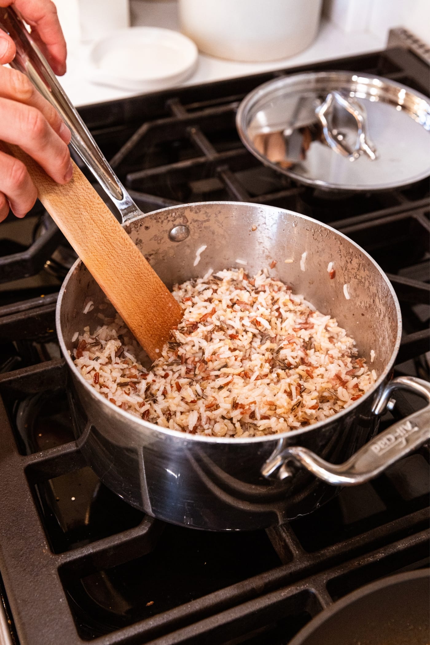 Hand holding wooden spatula stirring together wild rice in metal kettle on stovetop showing rice fully cooked.