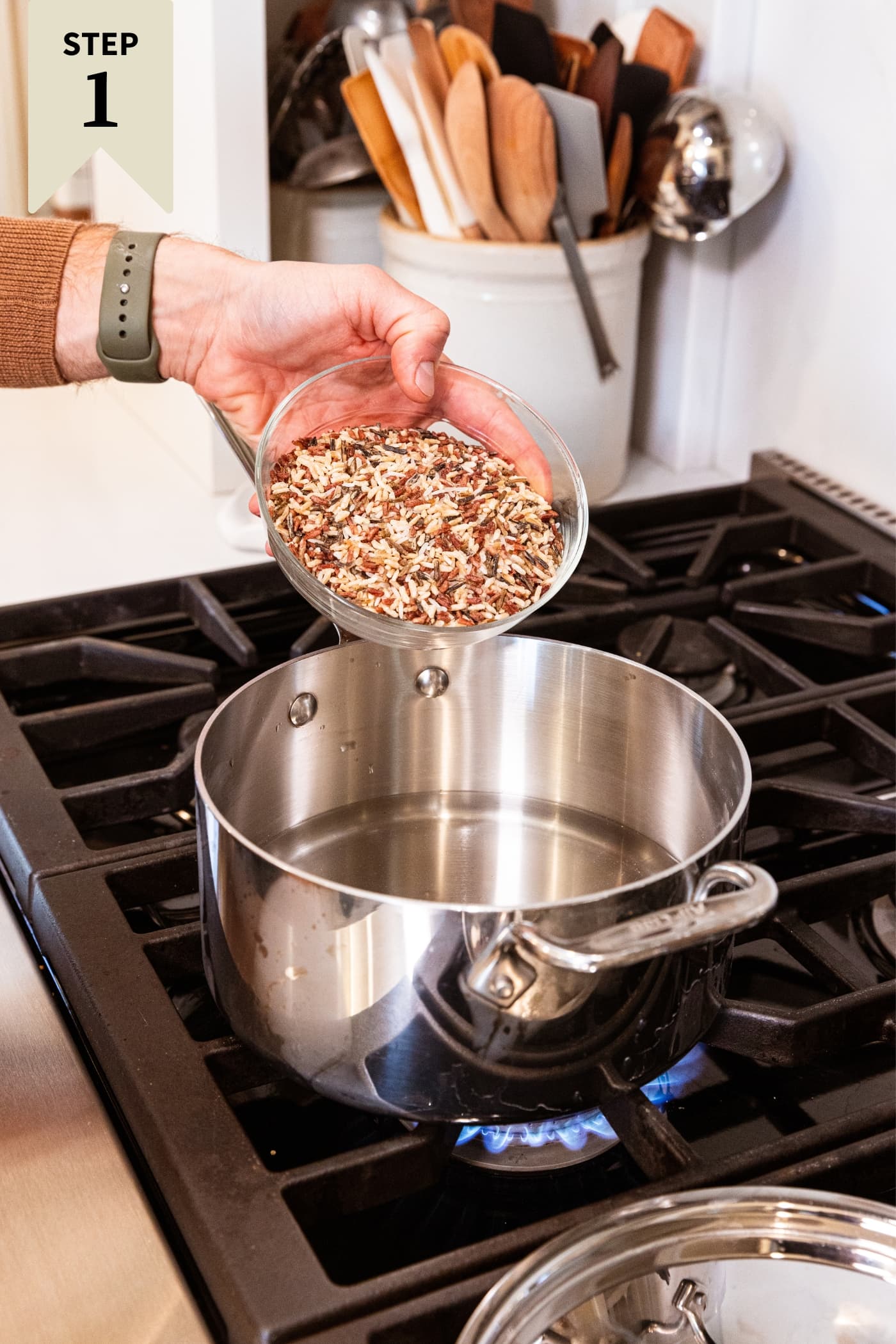 Hand pouring wild rice into a metal kettle filled with water sitting on stovetop.