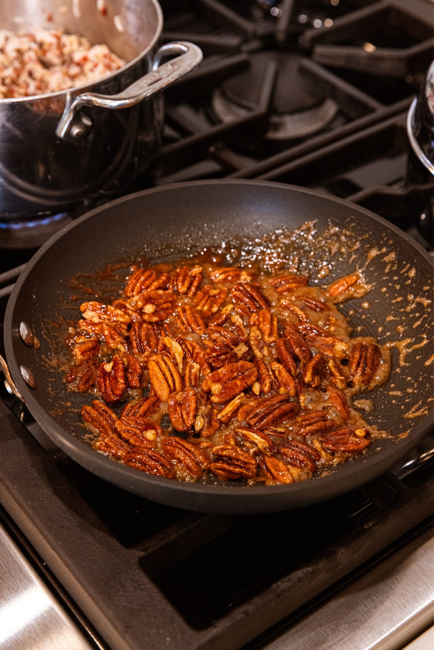 Small black skillet filled with pecans with melting brown sugar all around with small kettle in background filled with cooked wild rice.