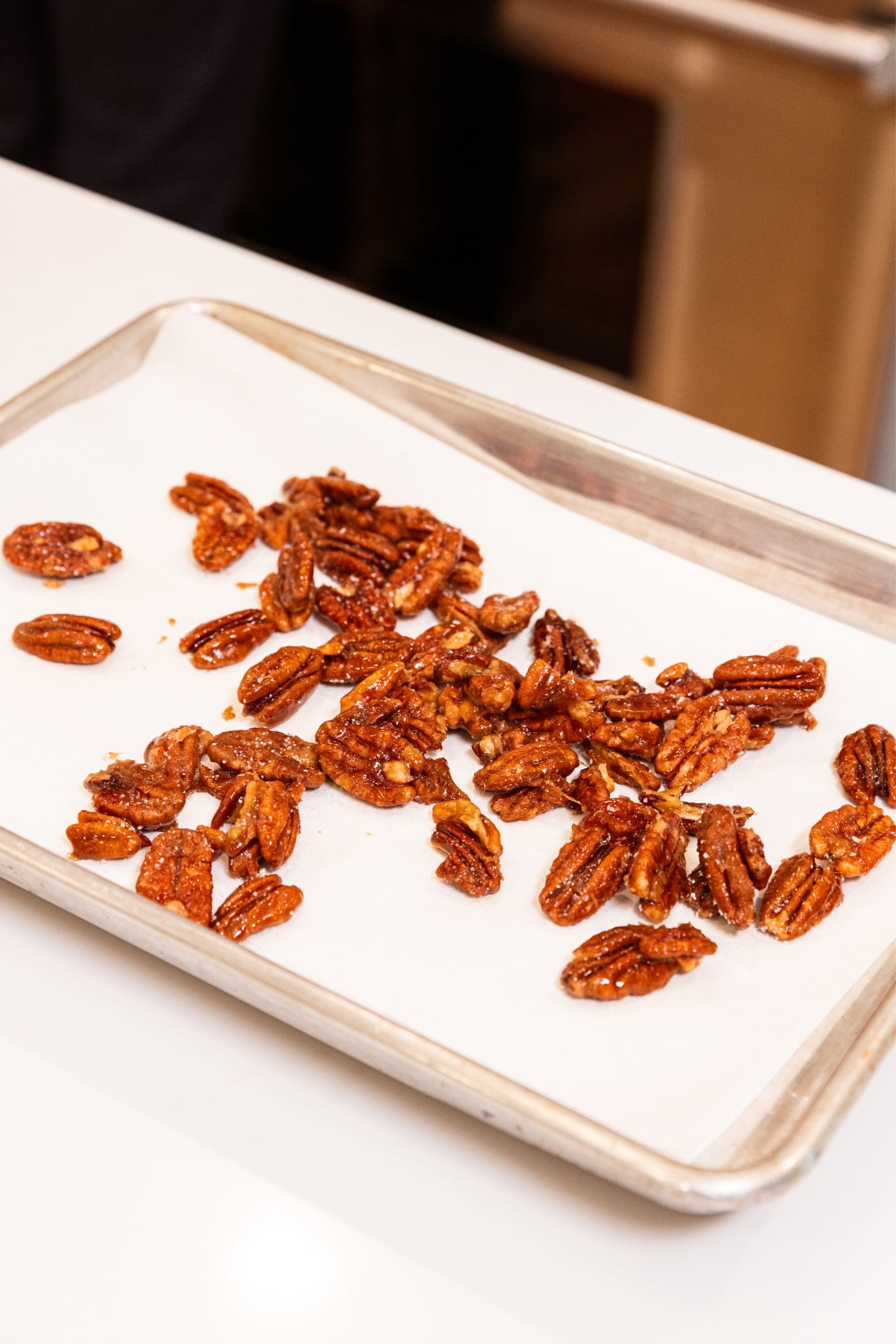 Praline candied pecans cooling on parchment-lined baking sheet sitting on white countertop with stove in background.