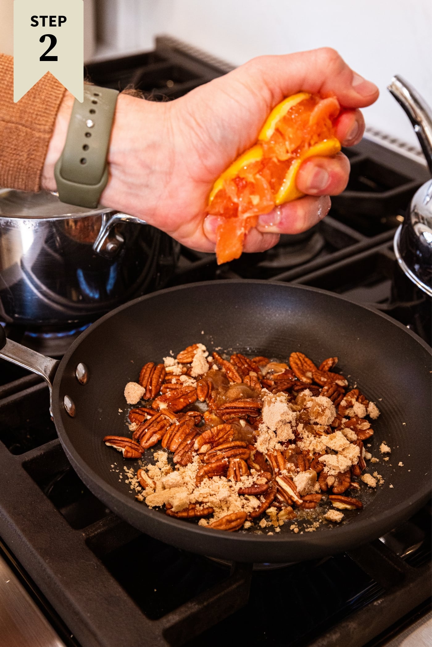 Hand squeezing orange over a small skillet filled with brown sugar and pecans sitting on stovetop before melting together.