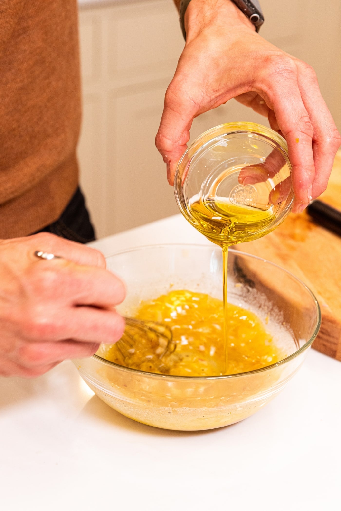 Hand holding small glass bowl pouring olive oil into larger glass bowl filled with ingredients needed to make homemade salad dressing.