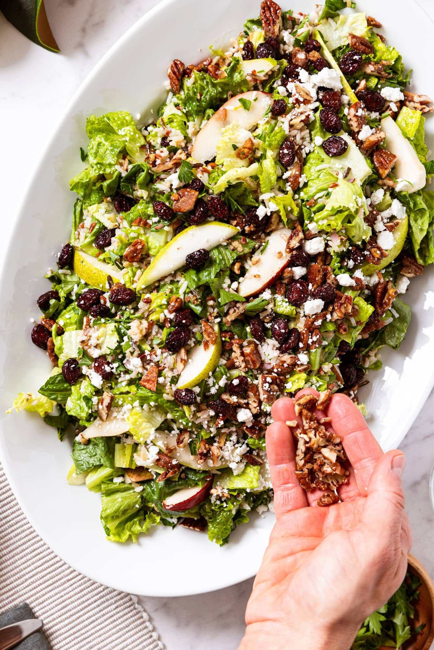 Hand holding chopped praline pecans over a large white platter filled with wild rice and pear salad topped with dried cranberries and feta cheese.
