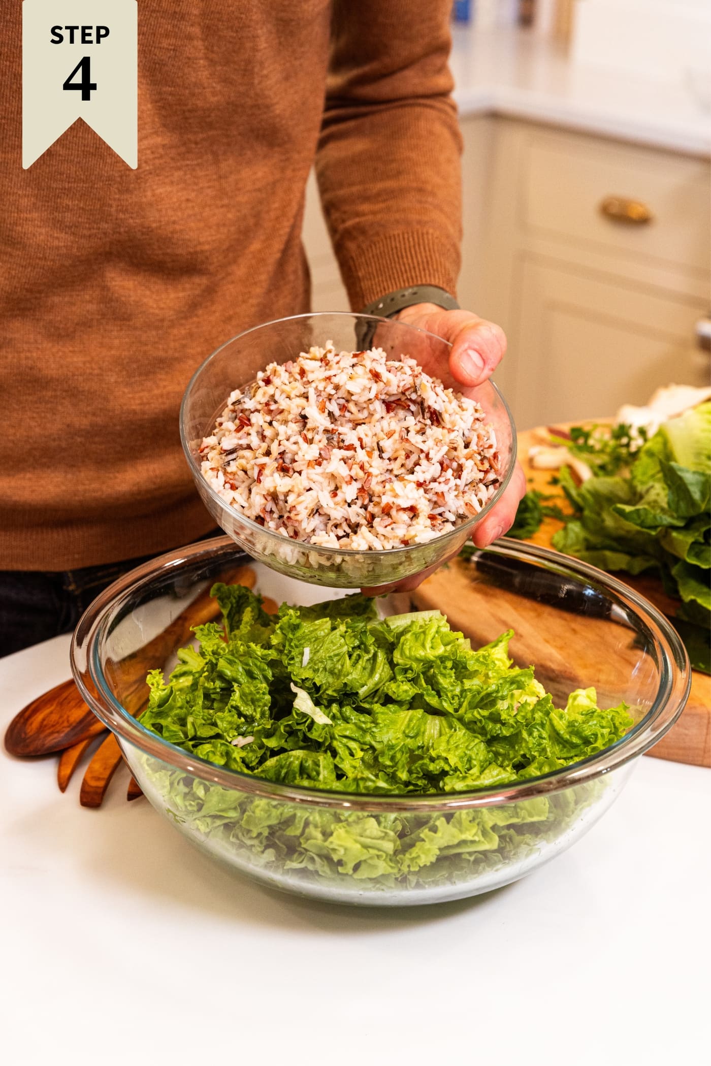 Hand holding small glass bowl filled with cooked wild rice about to pour contents into larger glass bowl filled with chopped lettuce with wood cutting board in background.