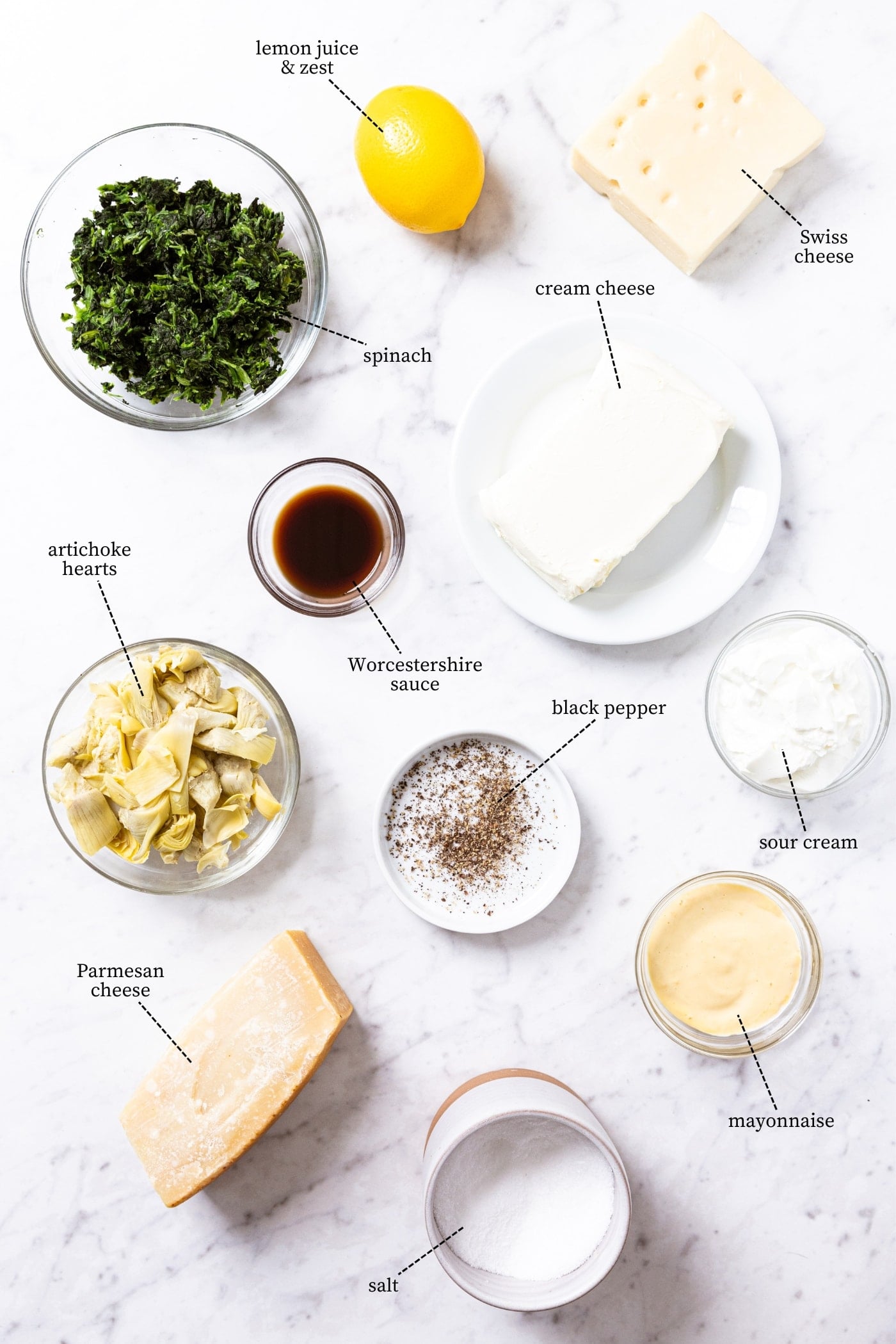 White marble surface filled with all of the ingredients needed to prepare baked spinach artichoke dip including spinach, artichoke hearts, cream cheese, sour cream, other cheeses, and more.