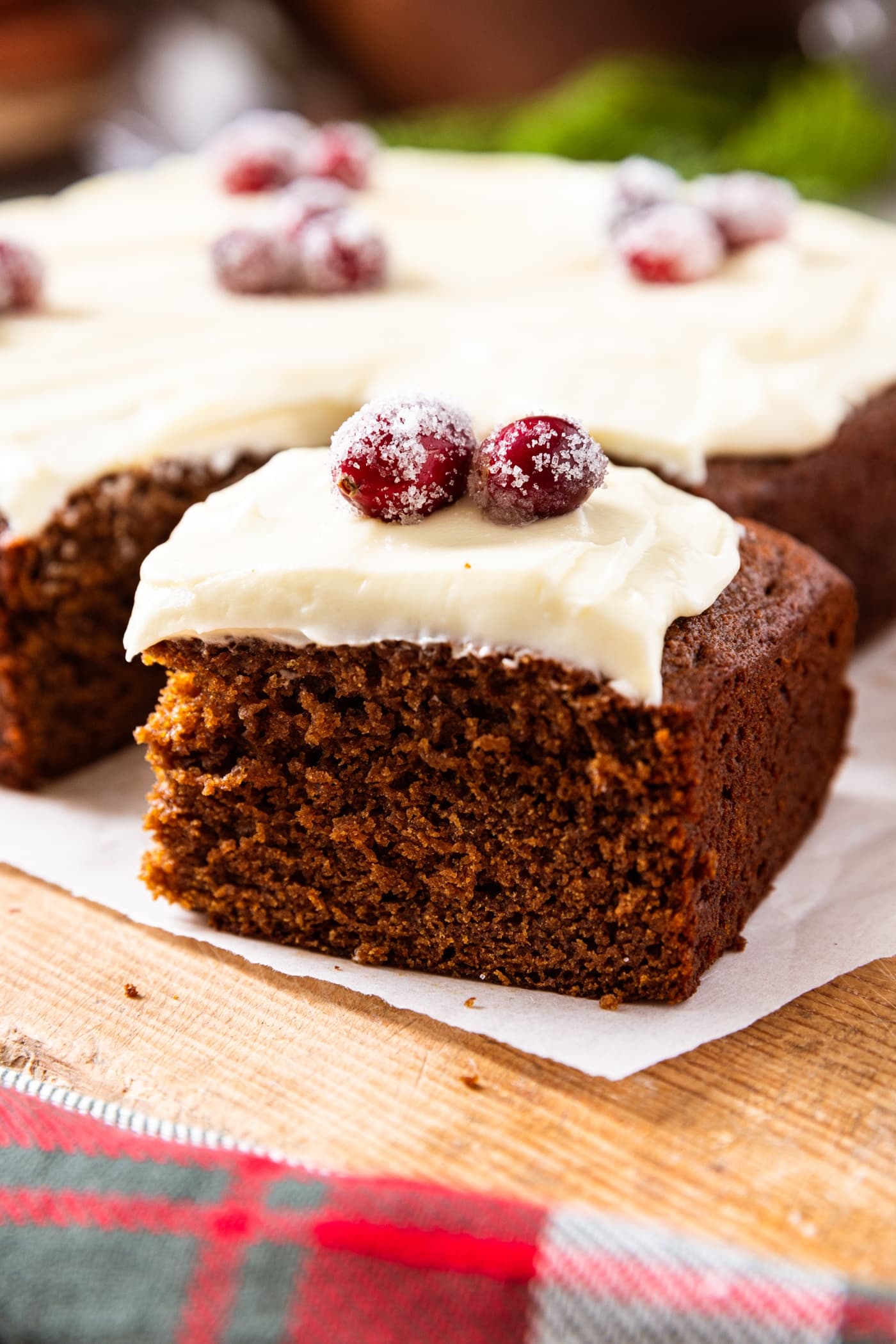 Square of gingerbread spice cake topped with cream cheese frosting and sugared cranberries sitting on piece of parchment on wood surface with the rest of the cake in the background.