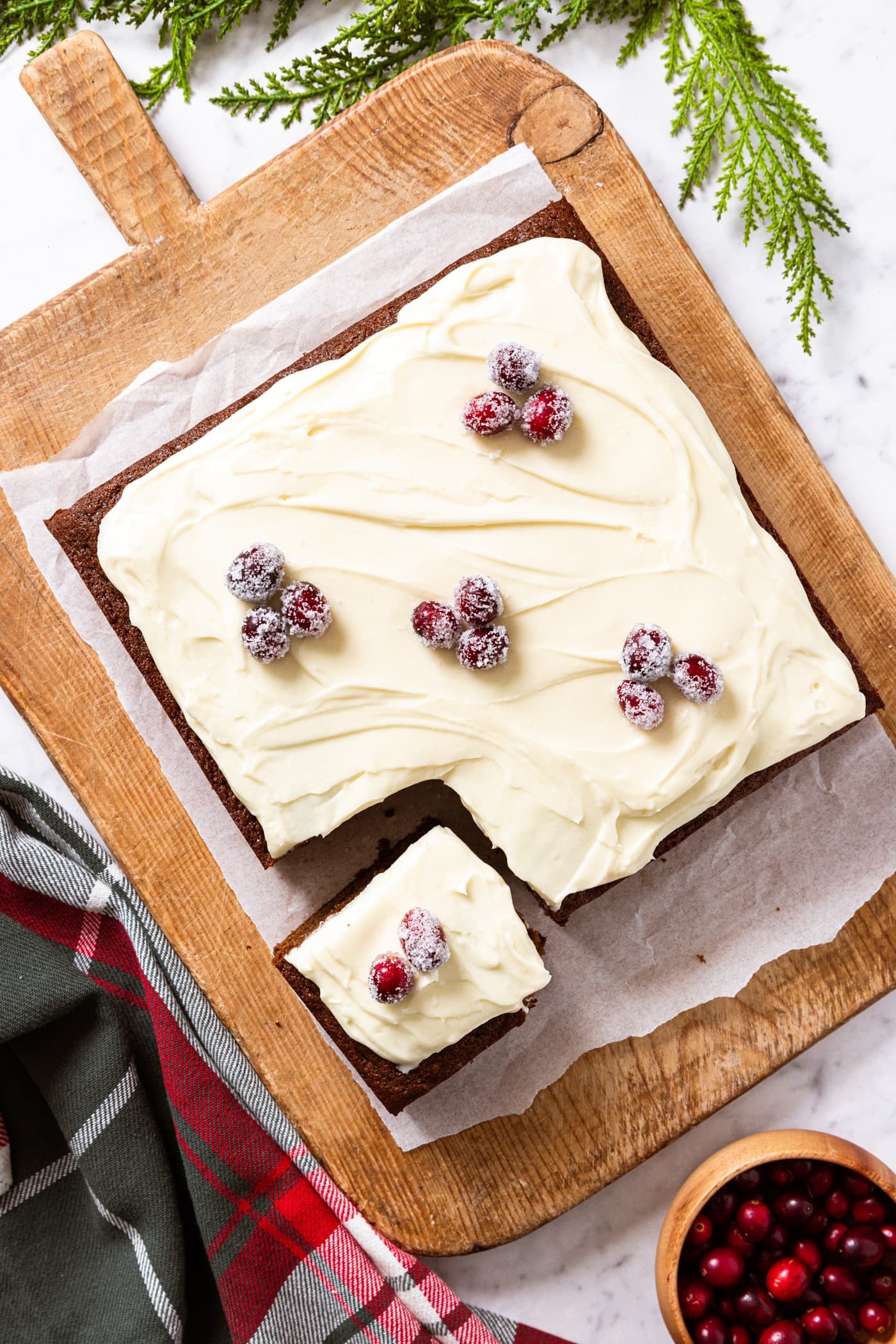 Wood cutting board with square gingerbread spice cake topped with sugared cranberries sitting on piece of white parchment all on marble surface with small bowl of cranberries and green garland.
