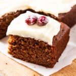 Square of gingerbread spice cake topped with cream cheese frosting and sugared cranberries sitting on piece of white parchment paper on wood cutting board with rest of the cake in the background.
