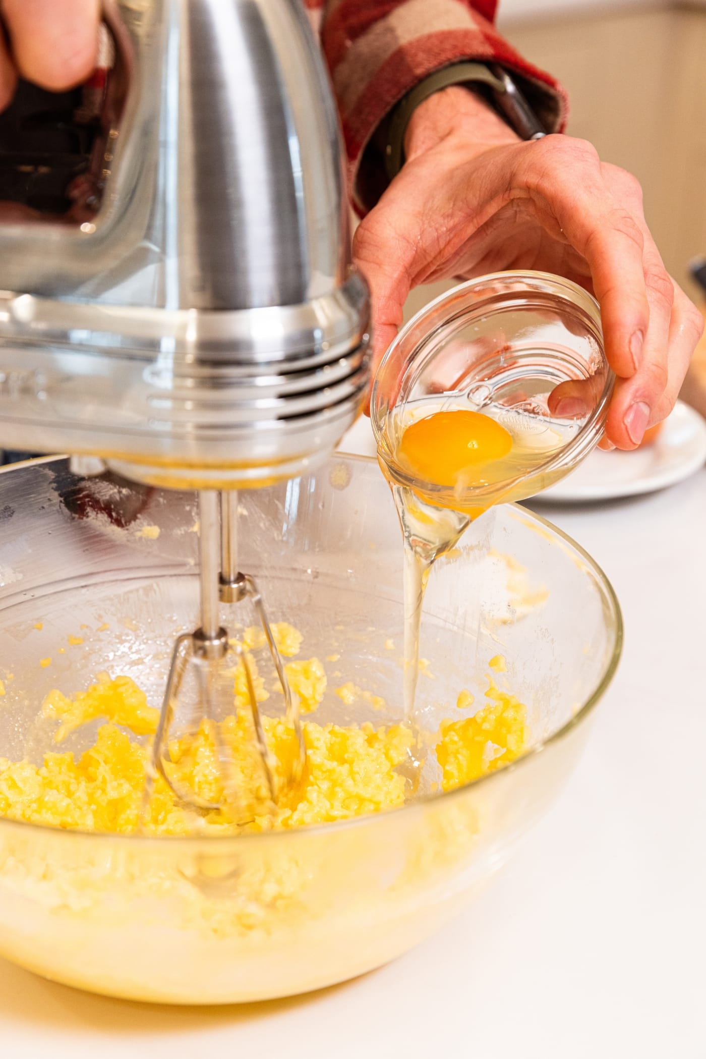 Hand holding small glass bowl with egg getting poured into a bowl filled with butter and sugar batter with hand mixer in it.