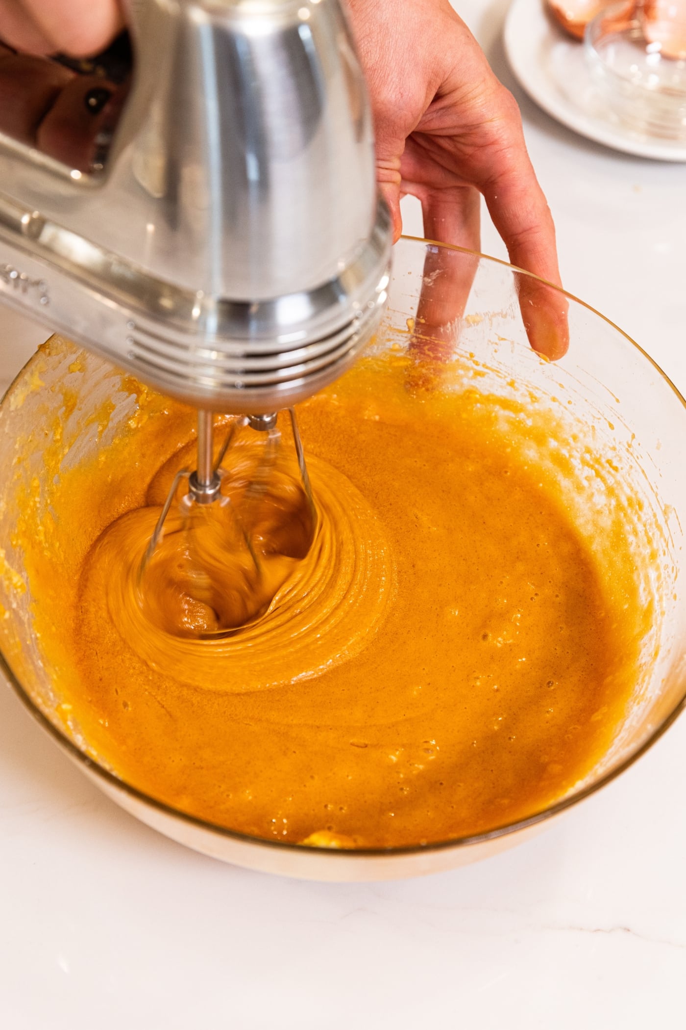 Glass mixing bowl filled with orange colored batter with a hand mixer actively mixing together the ingredients.