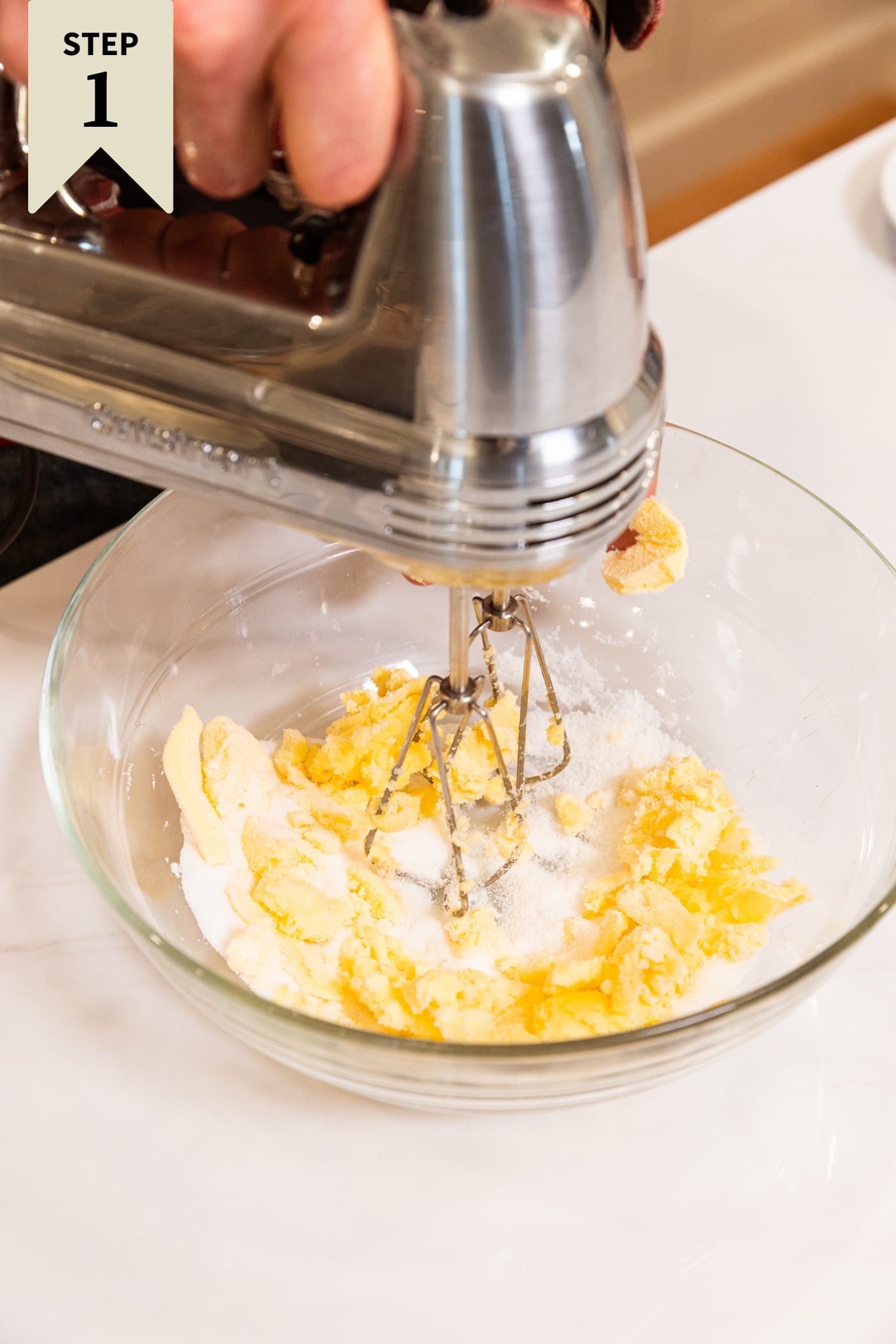 Glass mixing bowl filled with sugar and butter being mixed by hand mixer all on white countertop.