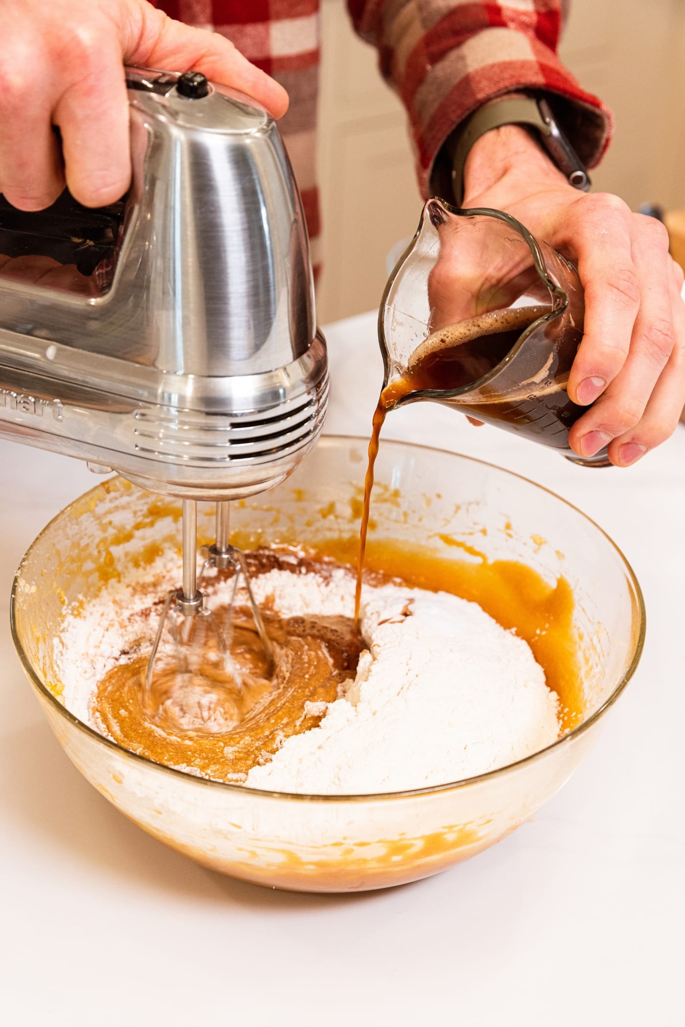 Hand holding hand mixer mixing together batter with dry ingredients while pouring brewed coffee into the mixture from a glass measuring cup.