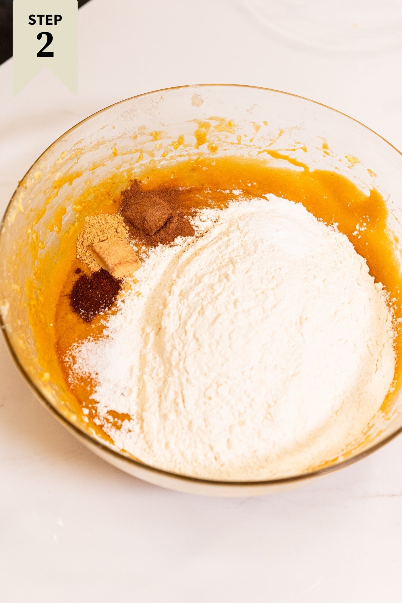 Glass mixing bowl filled with orange colored batter topped with flour and spices.