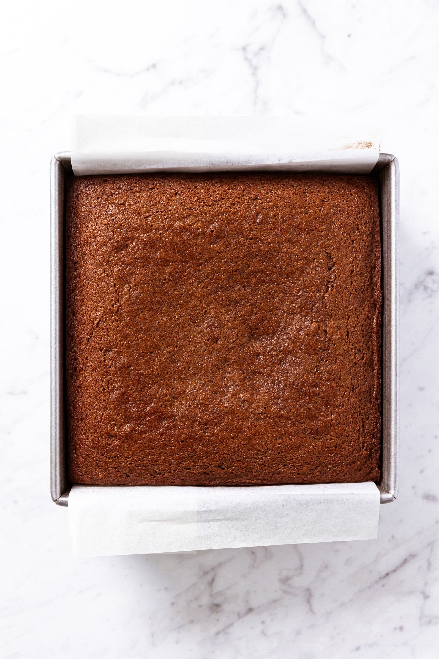 White marble surface with baked gingerbread spice cake with parchment underneath with browned top.