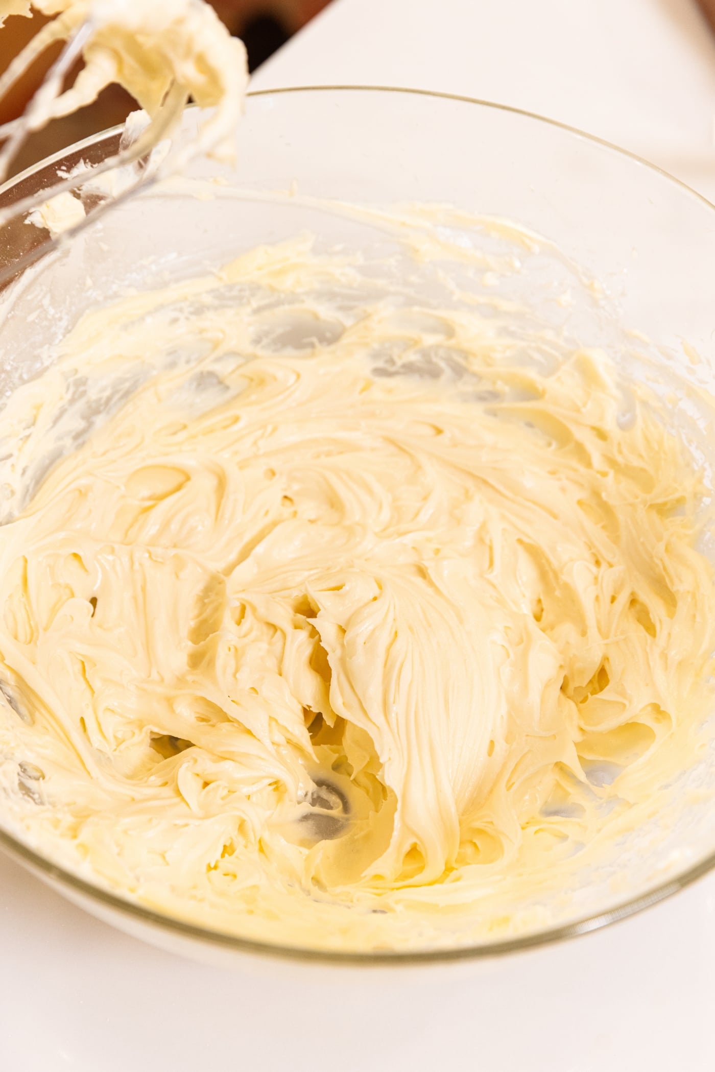 Lightly yellow colored cream cheese frosting in glass bowl after being mixed with hand mixer all on white countertop.