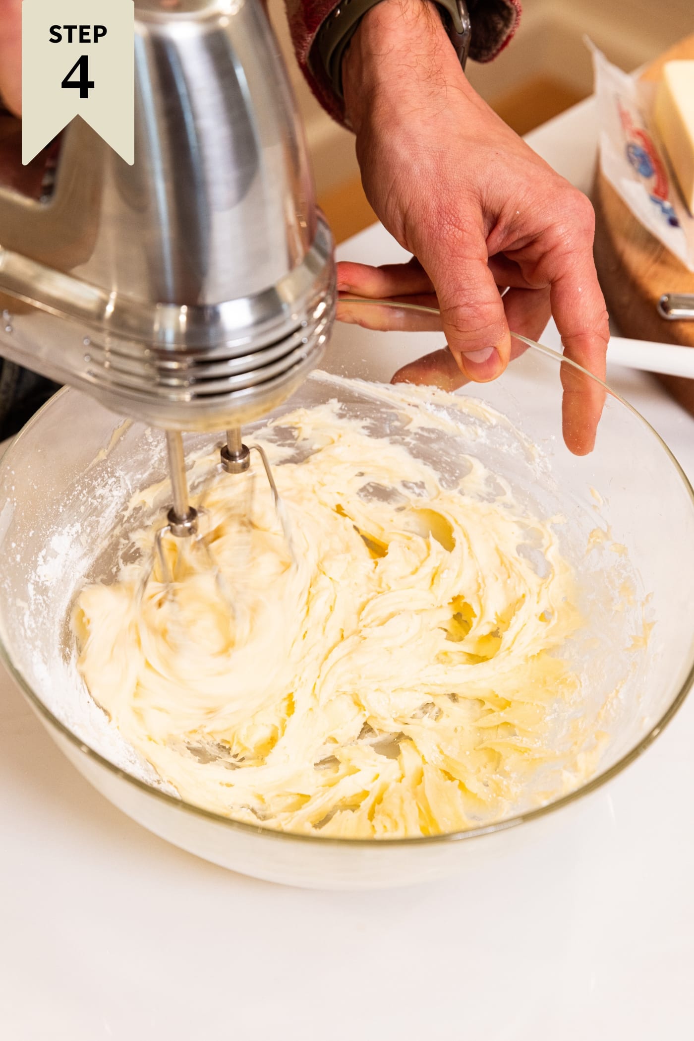 Glass bowl filled with cream cheese frosting being mixed with a hand mixer on white countertop.