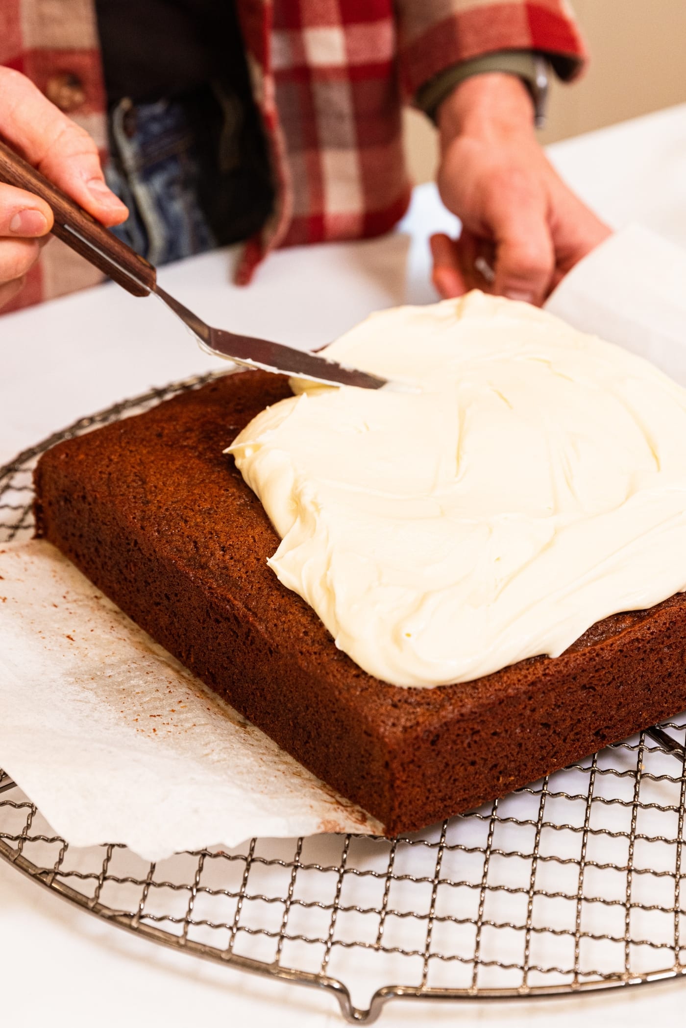 Hand using offset spatula to smooth out cream cheese frosting on top of gingerbread spice cake on round wire cooling rack.