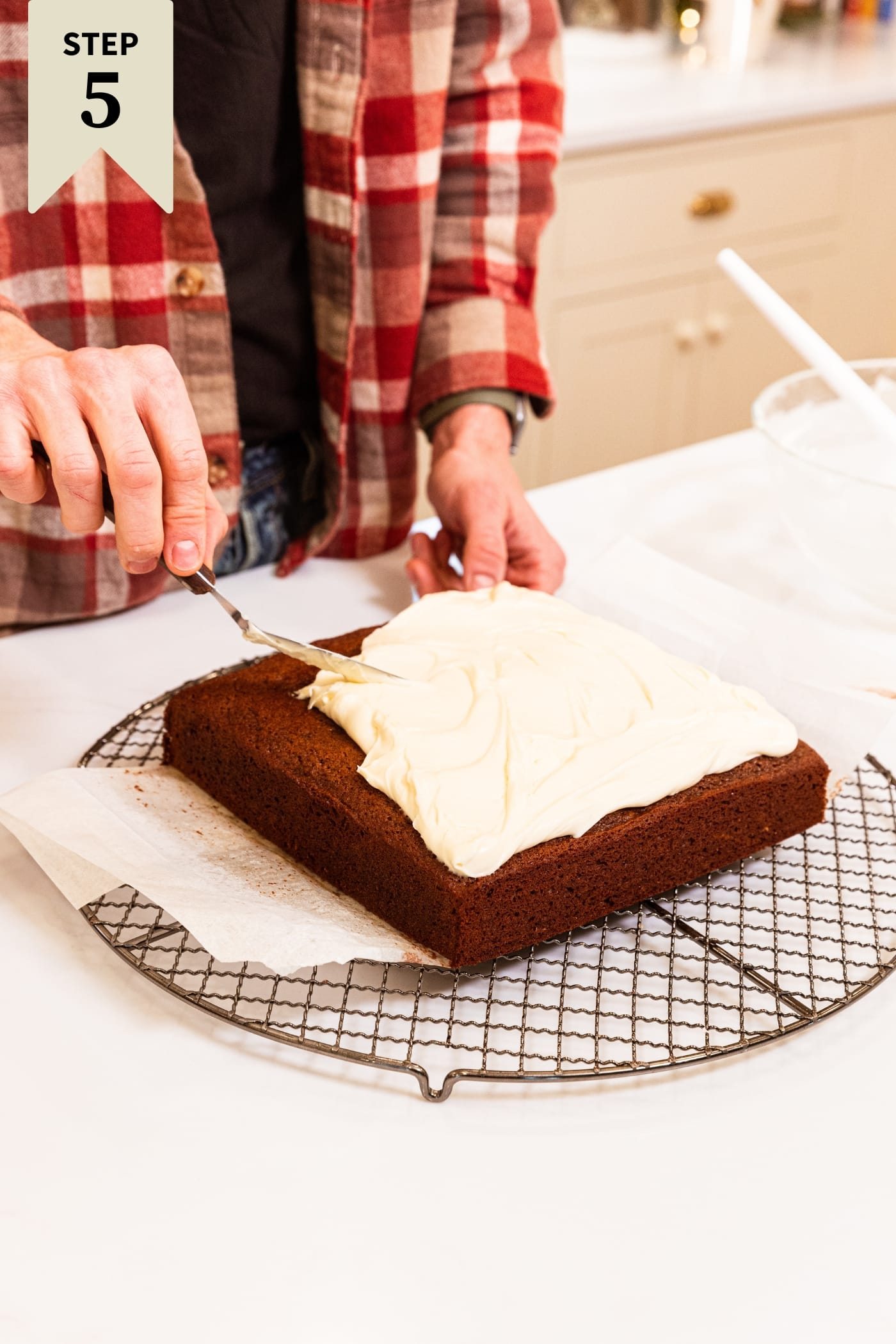 Hand spreading cream cheese frosting on top of a gingerbread spice cake sitting on round cooling rack with parchment underneath.