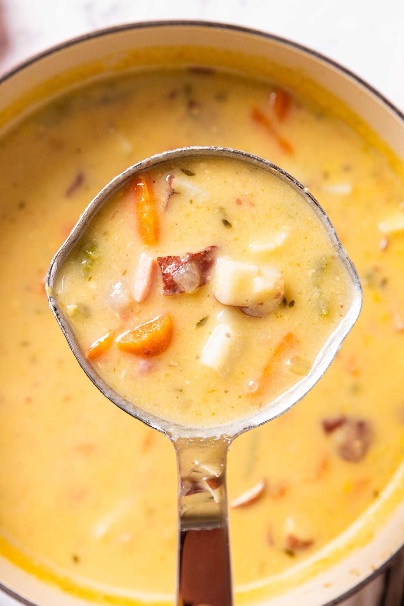 Ladle holding serving of ham and potato chowder over top of stockpot below holding the rest of the soup with pieces of carrot and ham showing through.