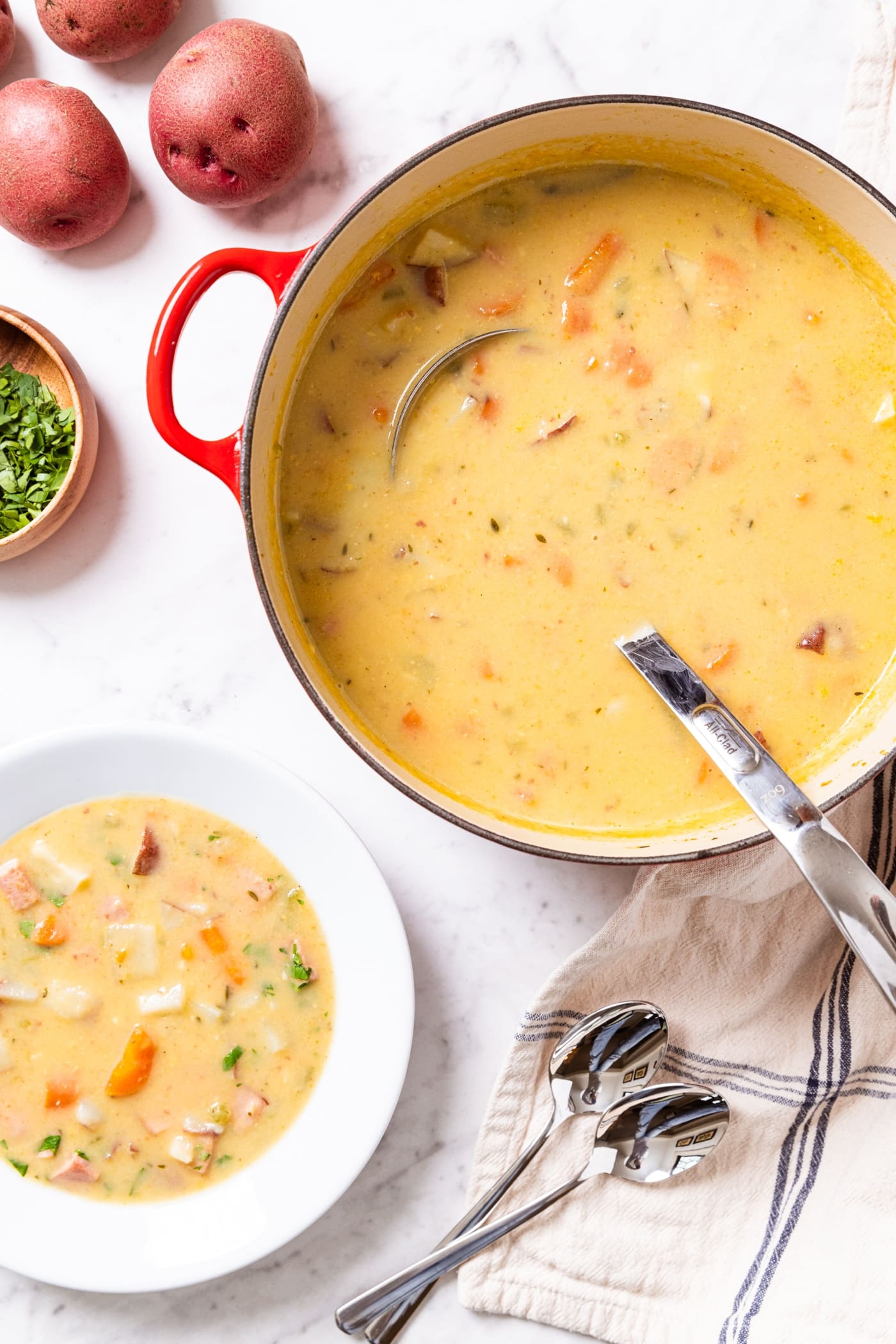 Large red stockpot filled with light yellow colored soup with white bowl to the side also filled with potato chowder with potatoes and parsley as well.