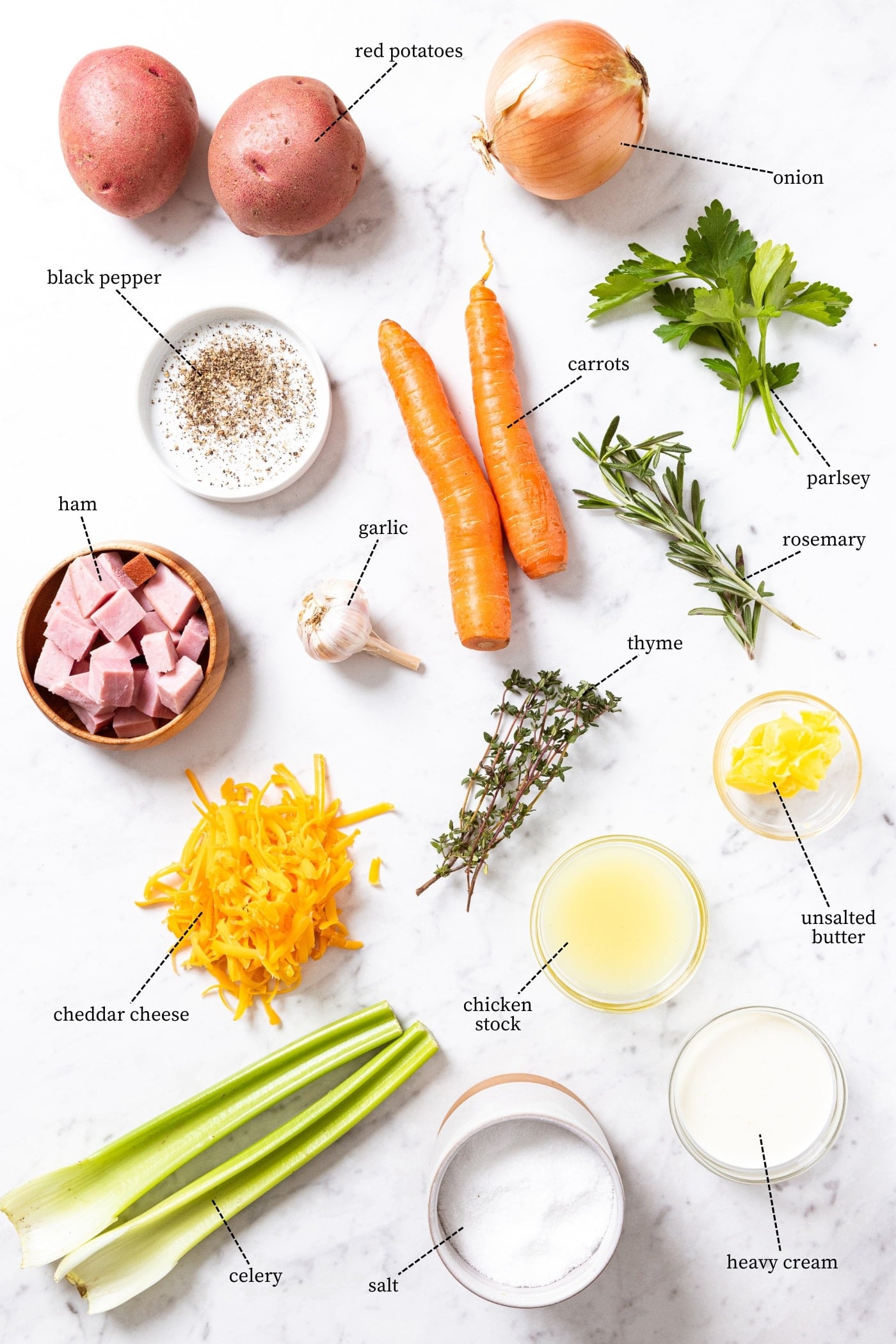 White marble surface filled with all ingredients needed to make ham and potato chowder including potatoes, ham, carrots, celery, cheese, herbs, spices, and more.