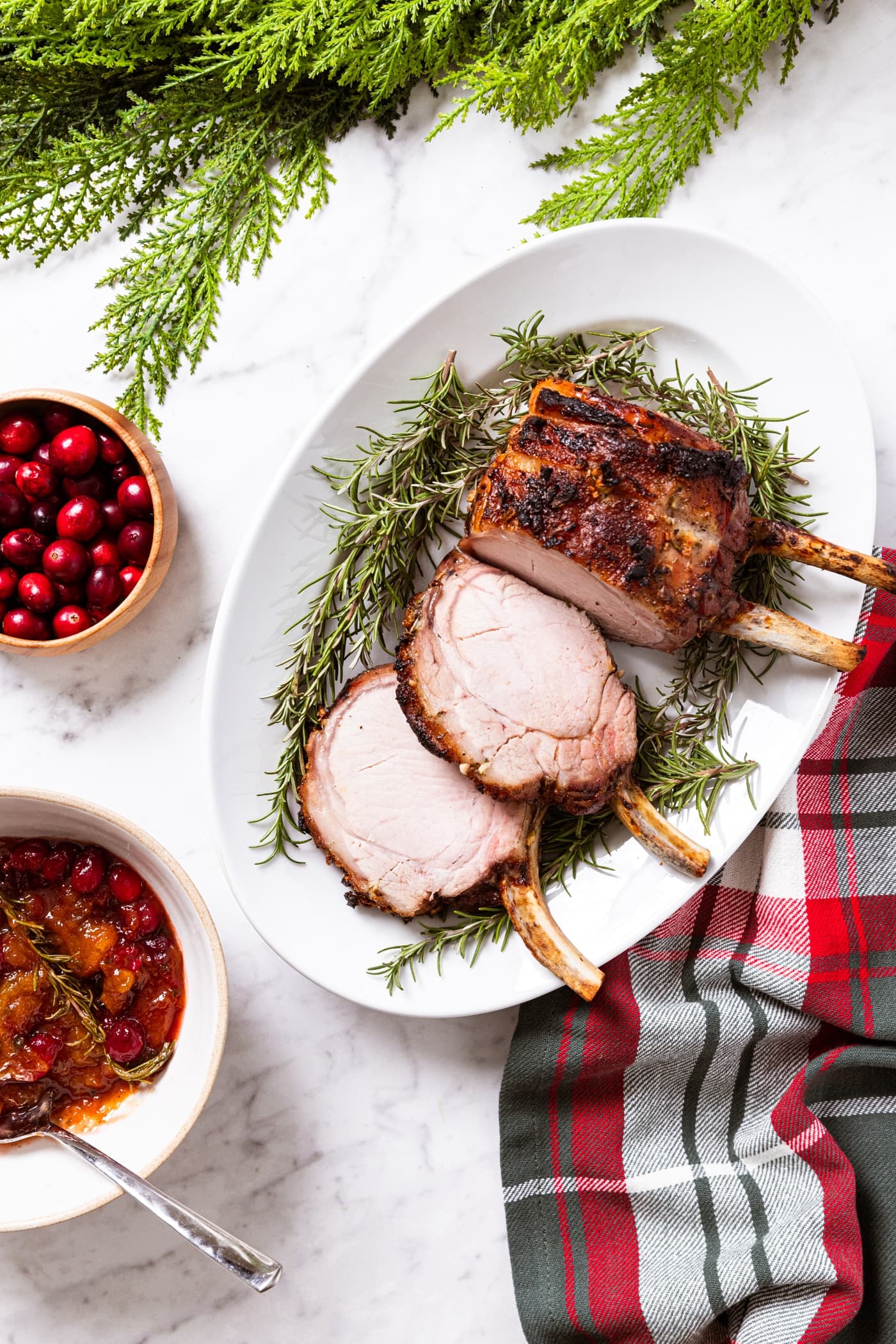 White oval platter with pork rib roast after being baked in the oven sitting on white marble countertop with cranberries and cranberry sauce to the side along with green garland.