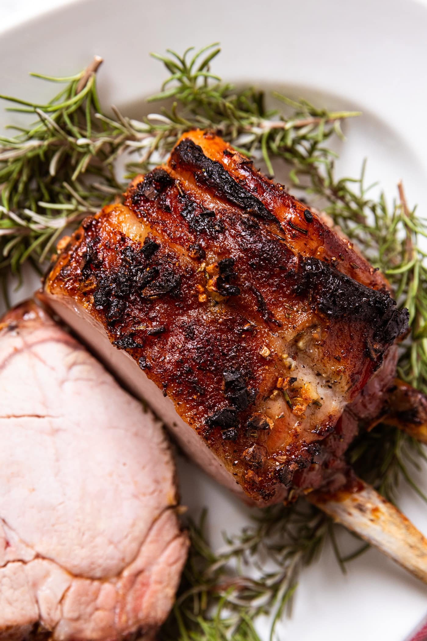 Crispy top texture of baked pork rib roast with slice sitting on to the side with fresh rosemary sprigs on the sides and underneath all on white oval platter.