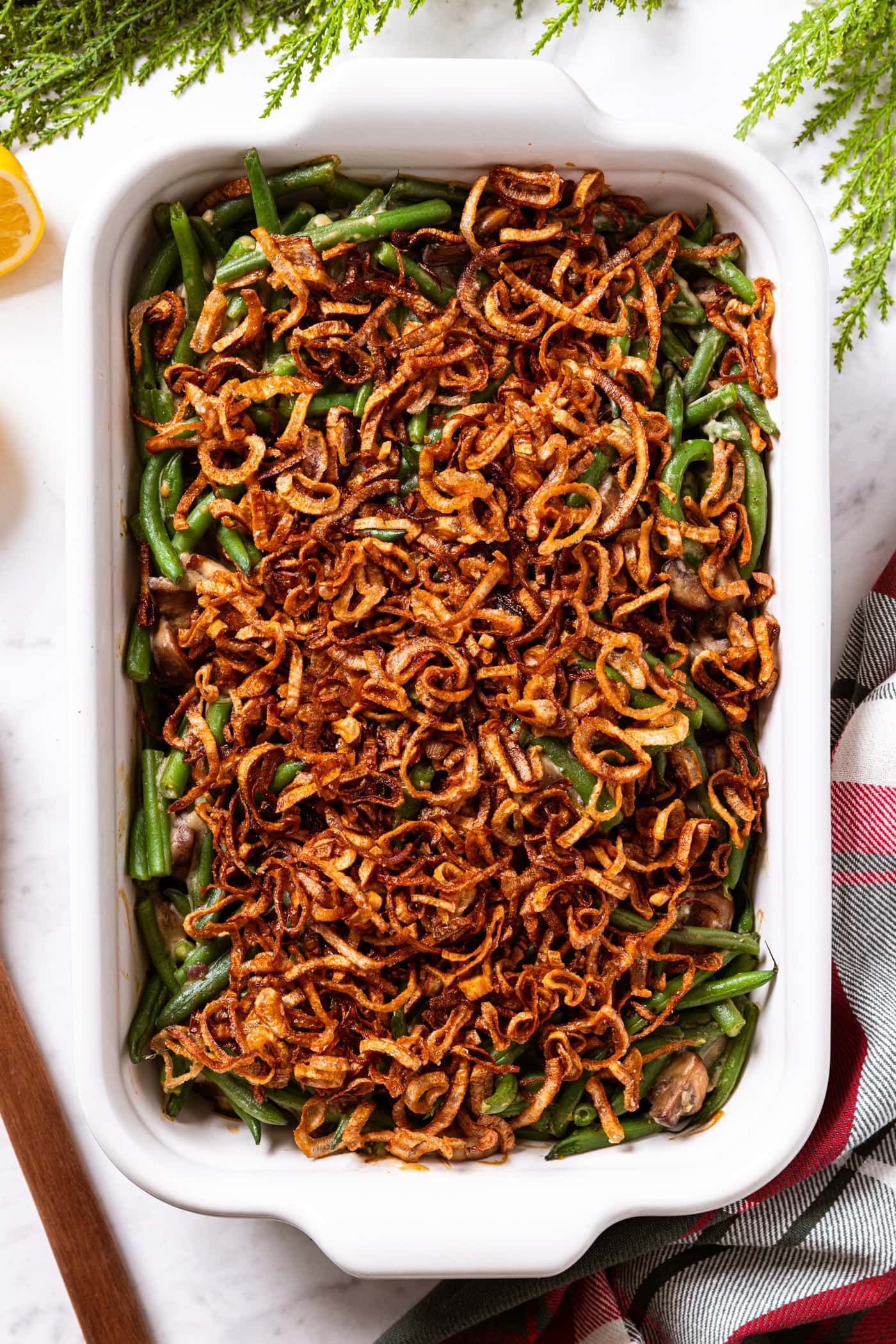 White baking dish filled with baked green bean casserole sitting on white marble surface with greenery all around as well as Christmas napkin topped with crispy fried shallots.