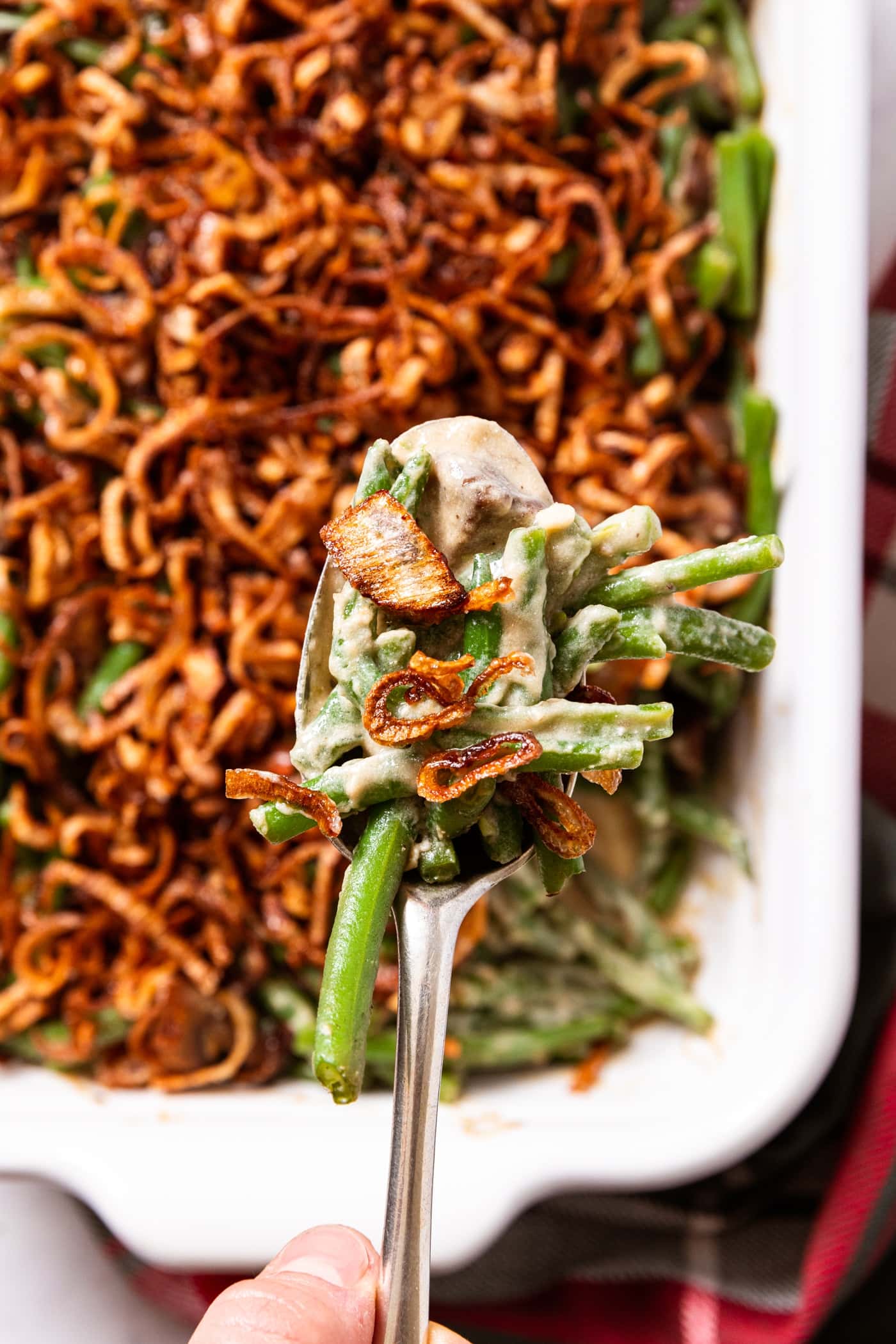 White baking dish filled with green beans with mushroom sauce with crispy fried shallots on top with hand holding spoonful after scooping from baking dish with Christmas napkin in front.