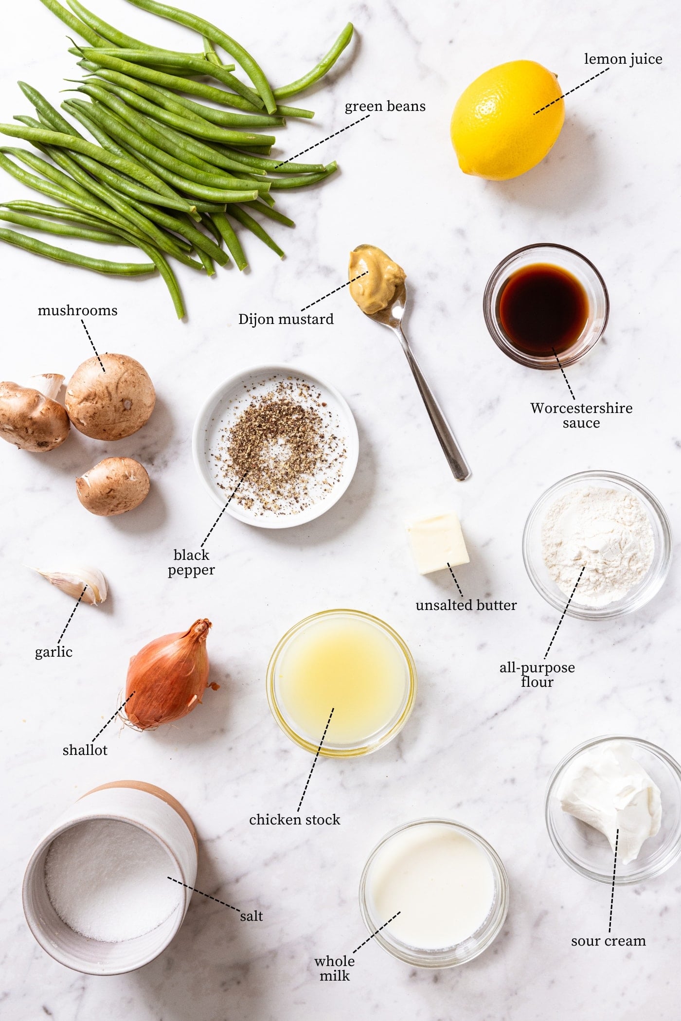 White marble surface filled with all ingredients needed to make green bean casserole including green beans, mushrooms, lemon, shallot, garlic, chicken stock, spices, and more.