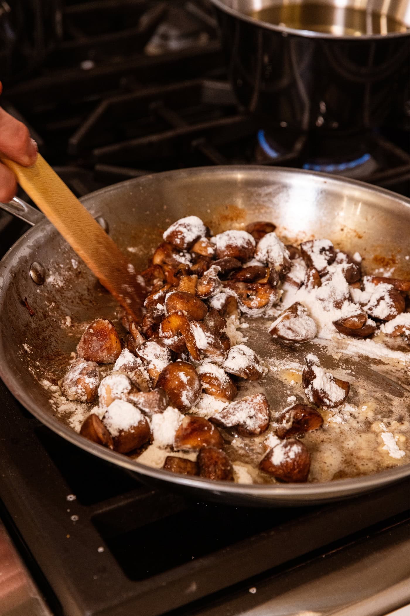 Cooking mushrooms being stirred with flour in a stainless steel pan on the stovetop.
