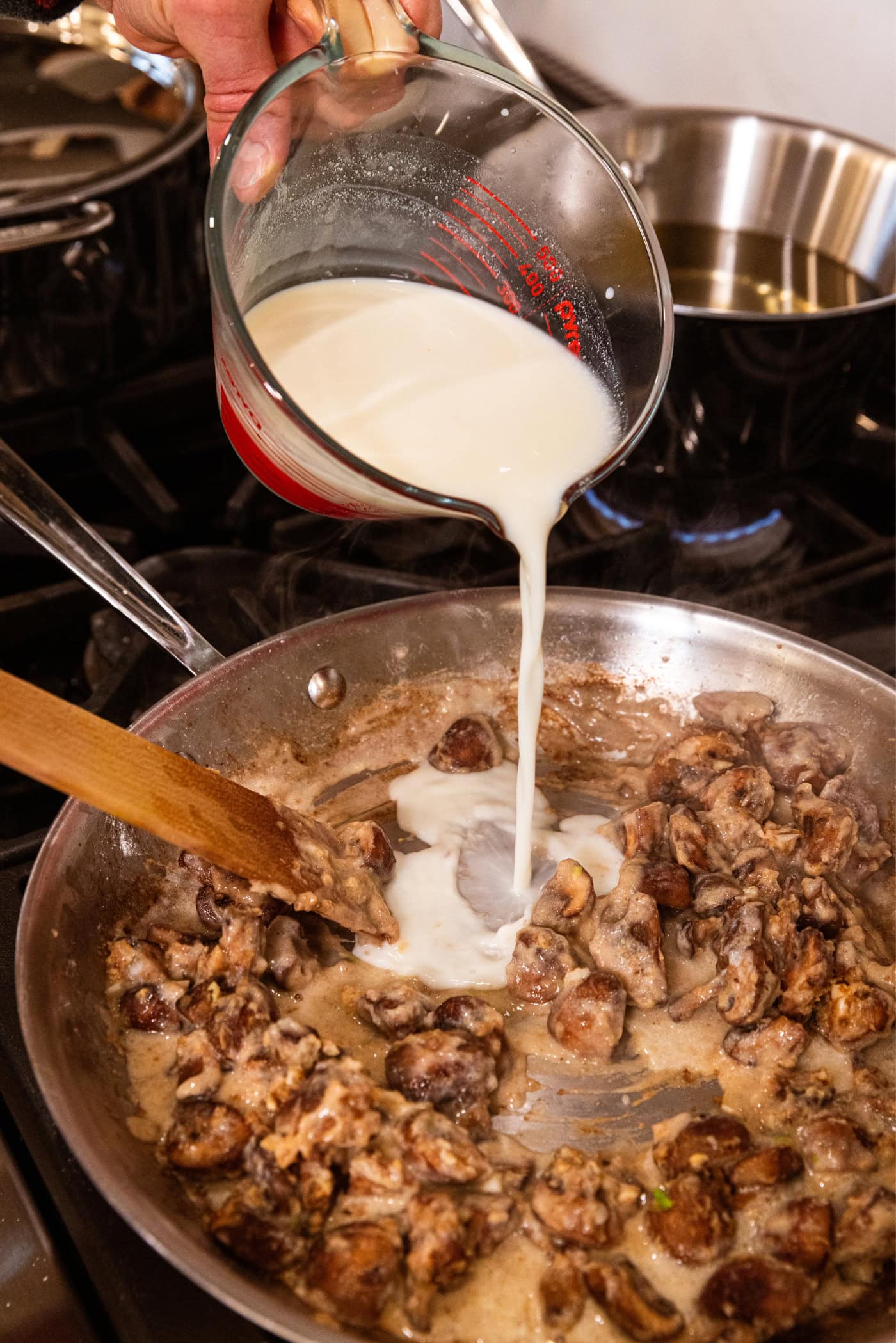 Milk pouring from a glass measuring cup into a pan filled with cooking mushrooms on the stovetop.