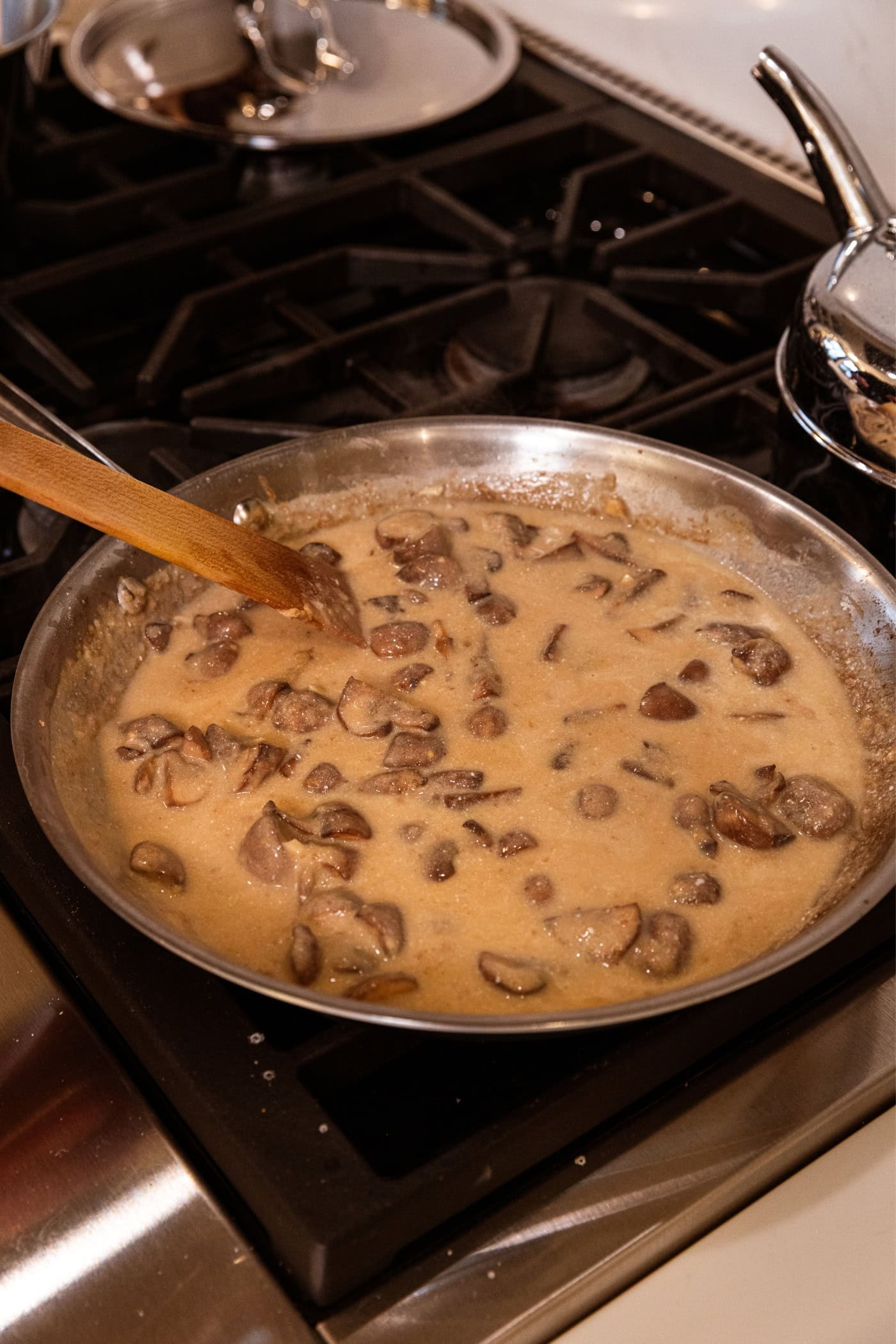 Thick mushroom sauce in a large stainless steel pan on the stovetop being stirred together with a wooden spatula.