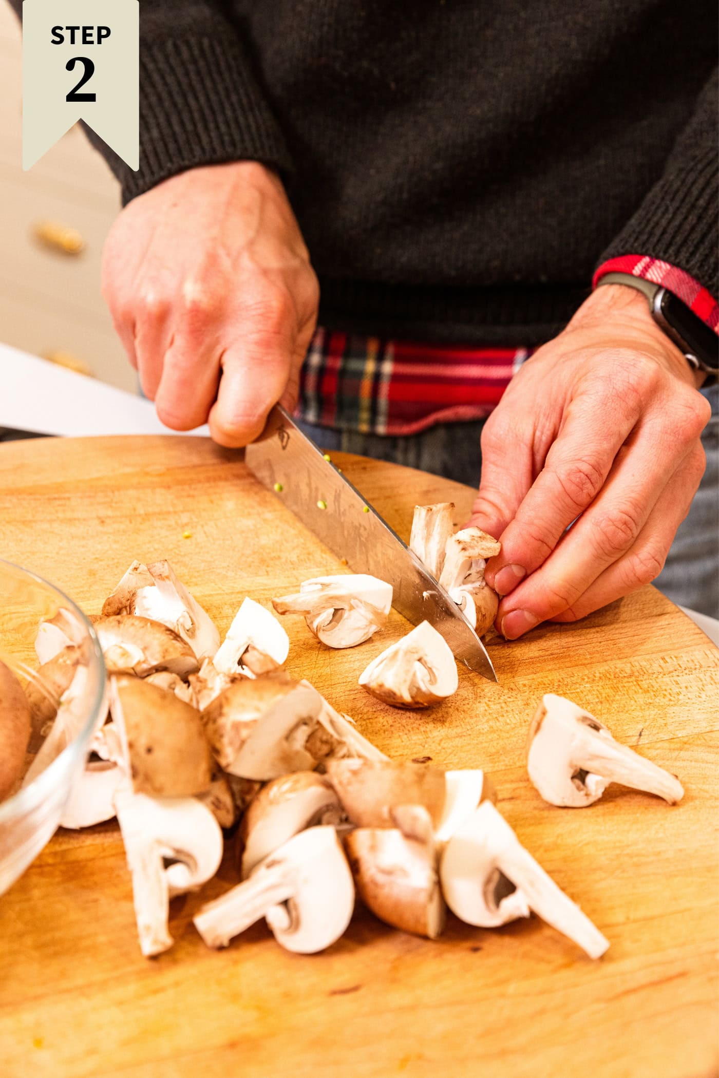 Hand using a knife to slice mushrooms into pieces on a wooden cutting board.