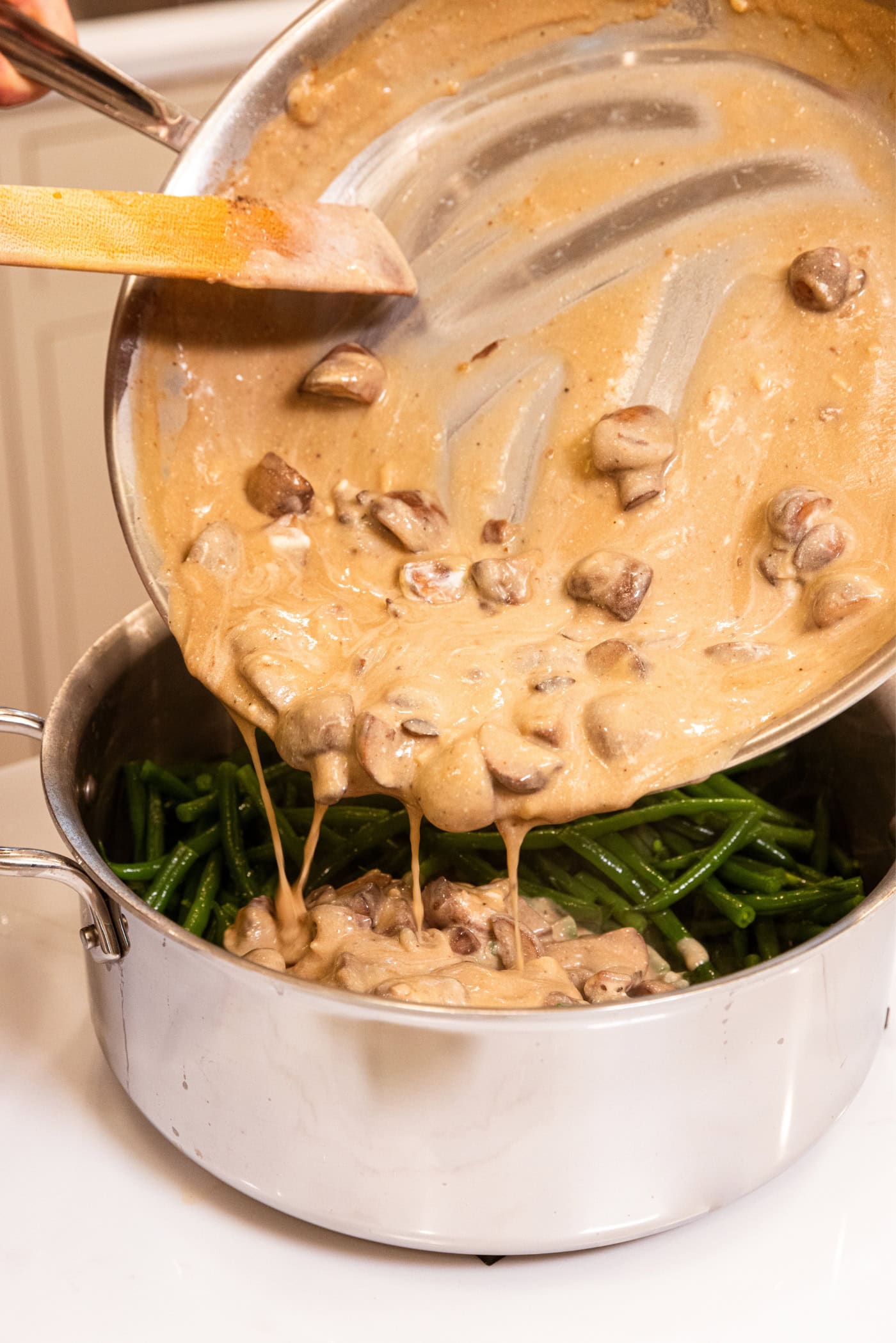 Green beans in a large stainless steel pot with a mushroom sauce being poured over the top.
